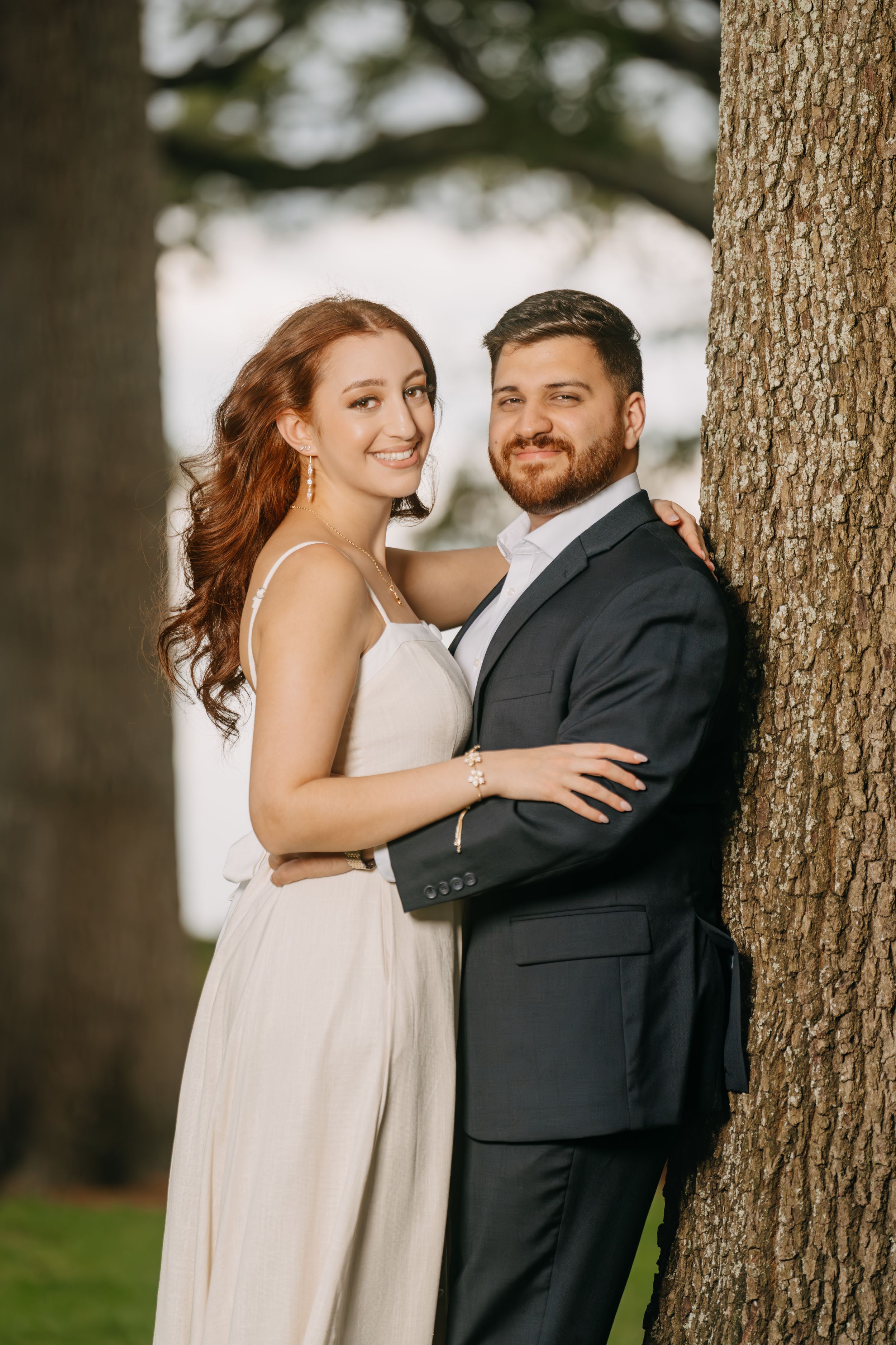 A smiling couple in formal attire poses near a large tree outdoors, the woman in a white dress and man in a dark suit.