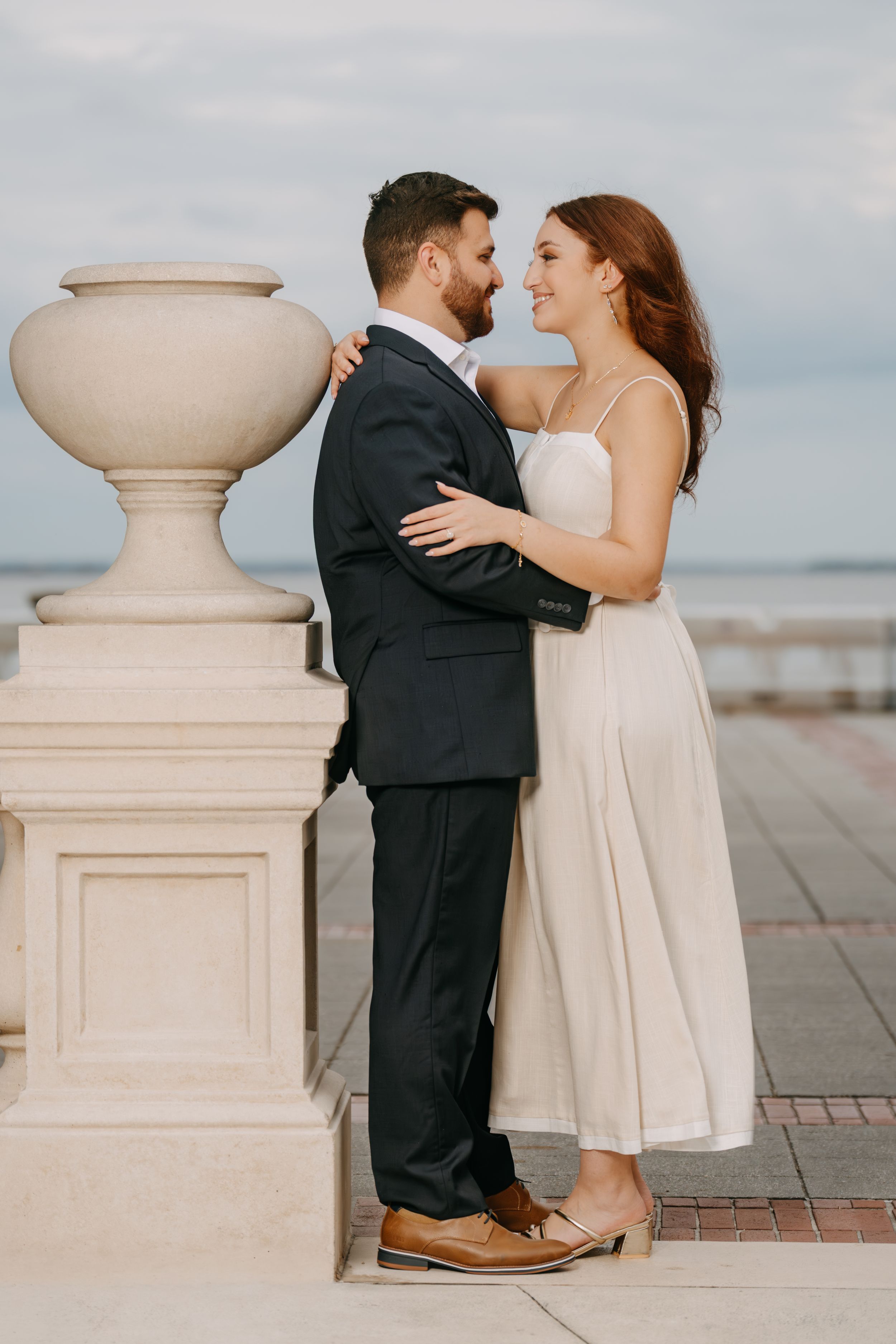 Couple in formal attire sharing a romantic moment on a waterfront terrace, woman in white dress embracing man in dark suit.