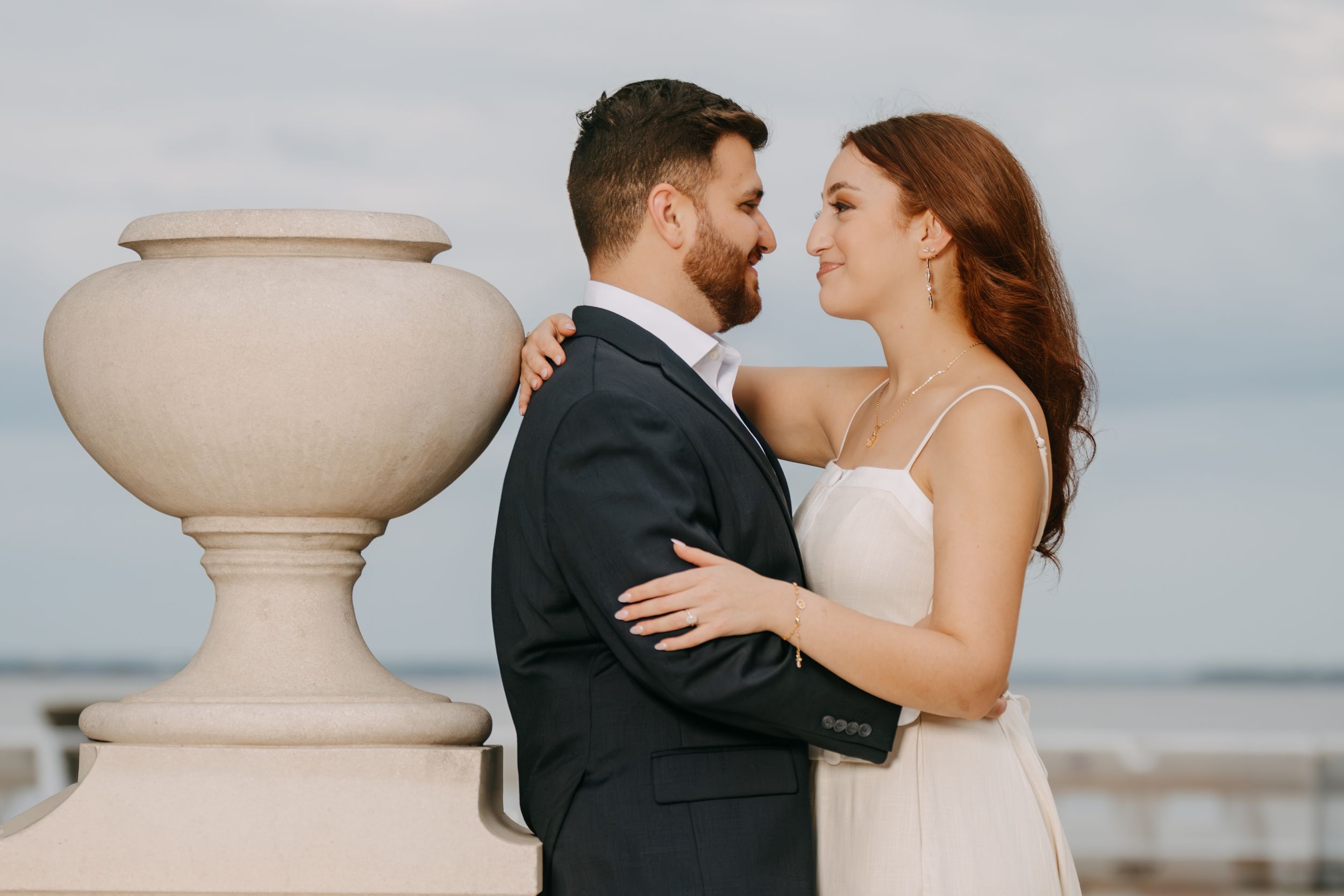 A bride and groom share a romantic moment near a stone balustrade with an ocean backdrop on their wedding day.