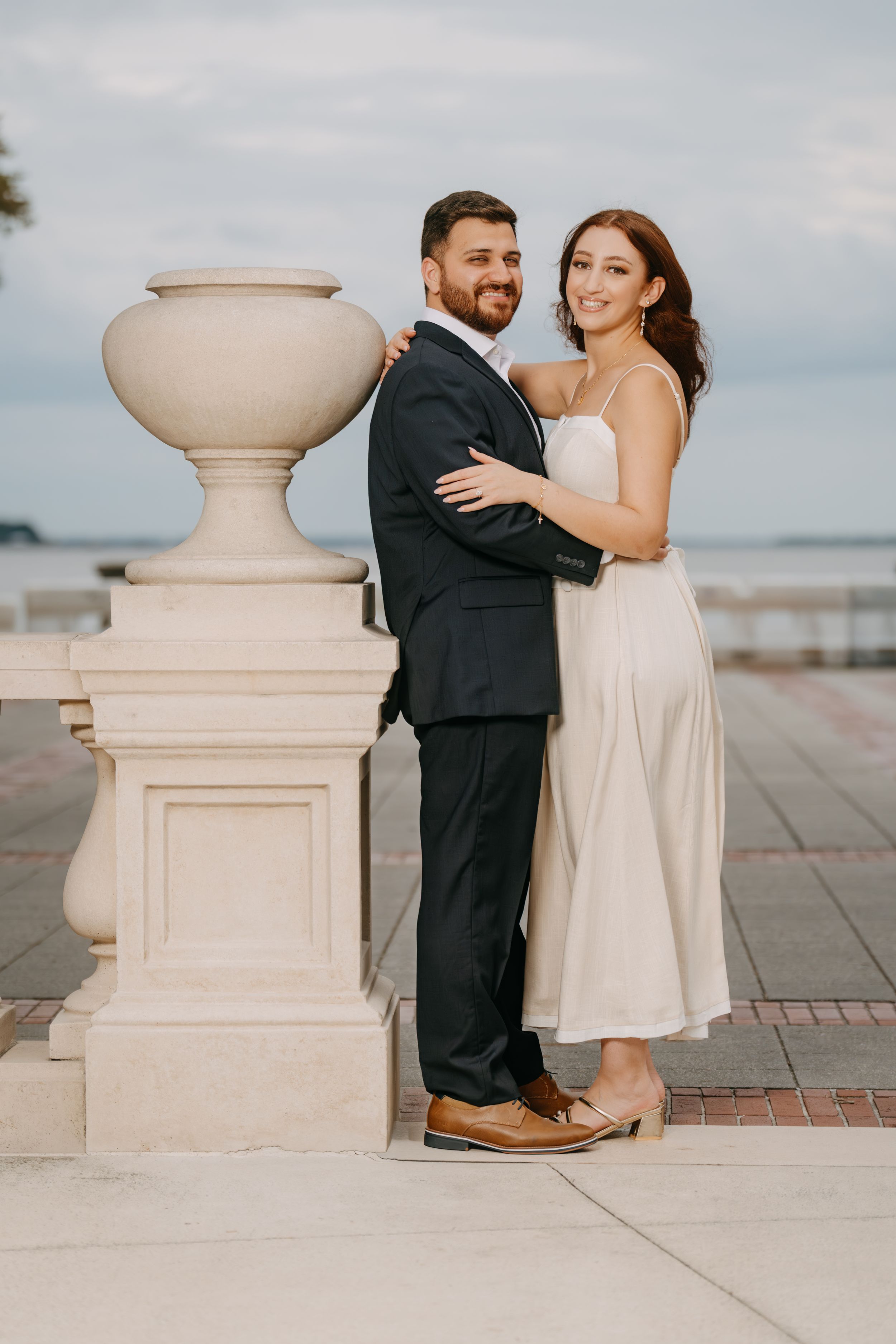 Couple embracing near ornate stone balustrade by waterfront, man in dark suit, woman in white midi dress.
