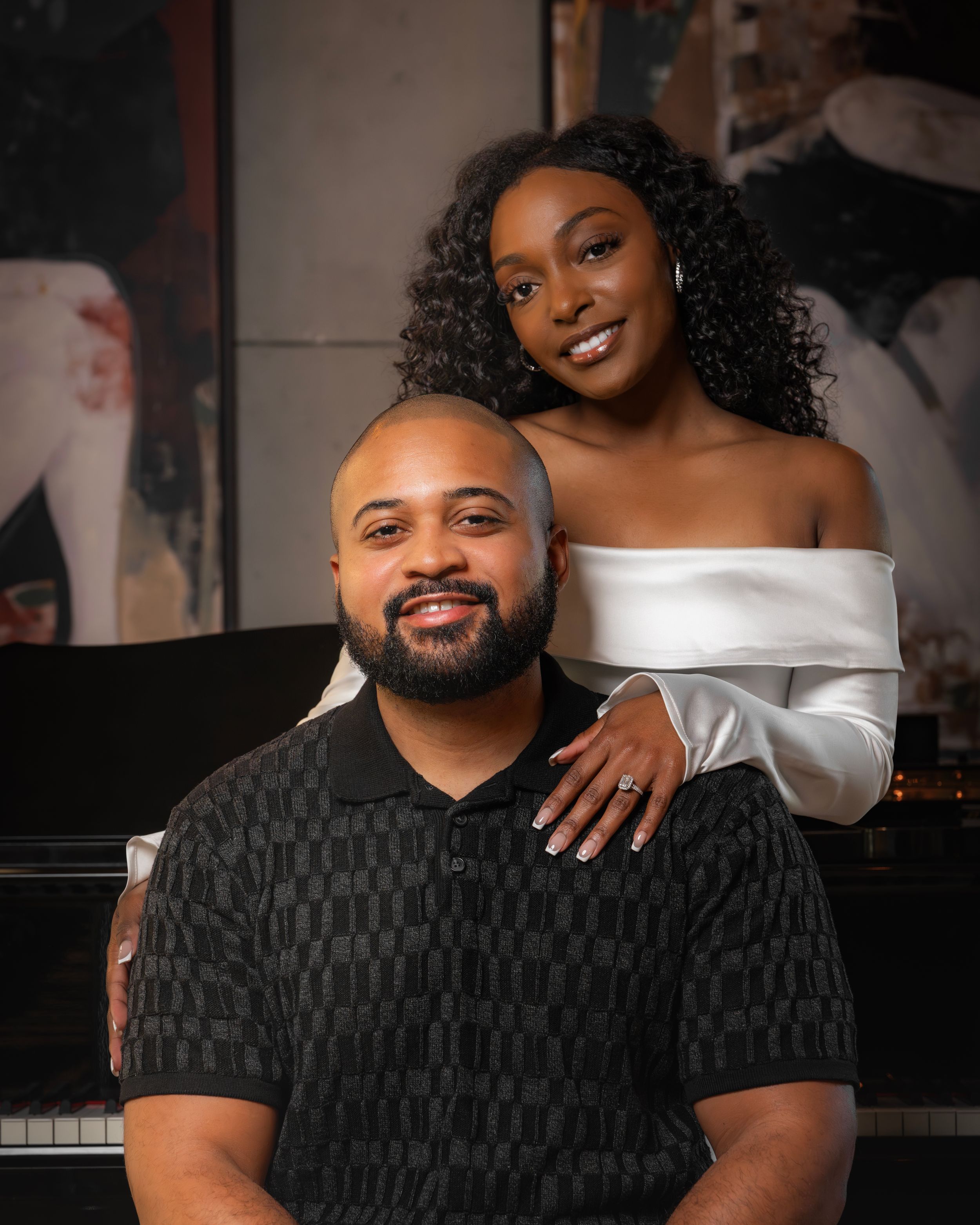 A smiling couple poses elegantly, woman in white off-shoulder top standing behind seated man in black patterned shirt.