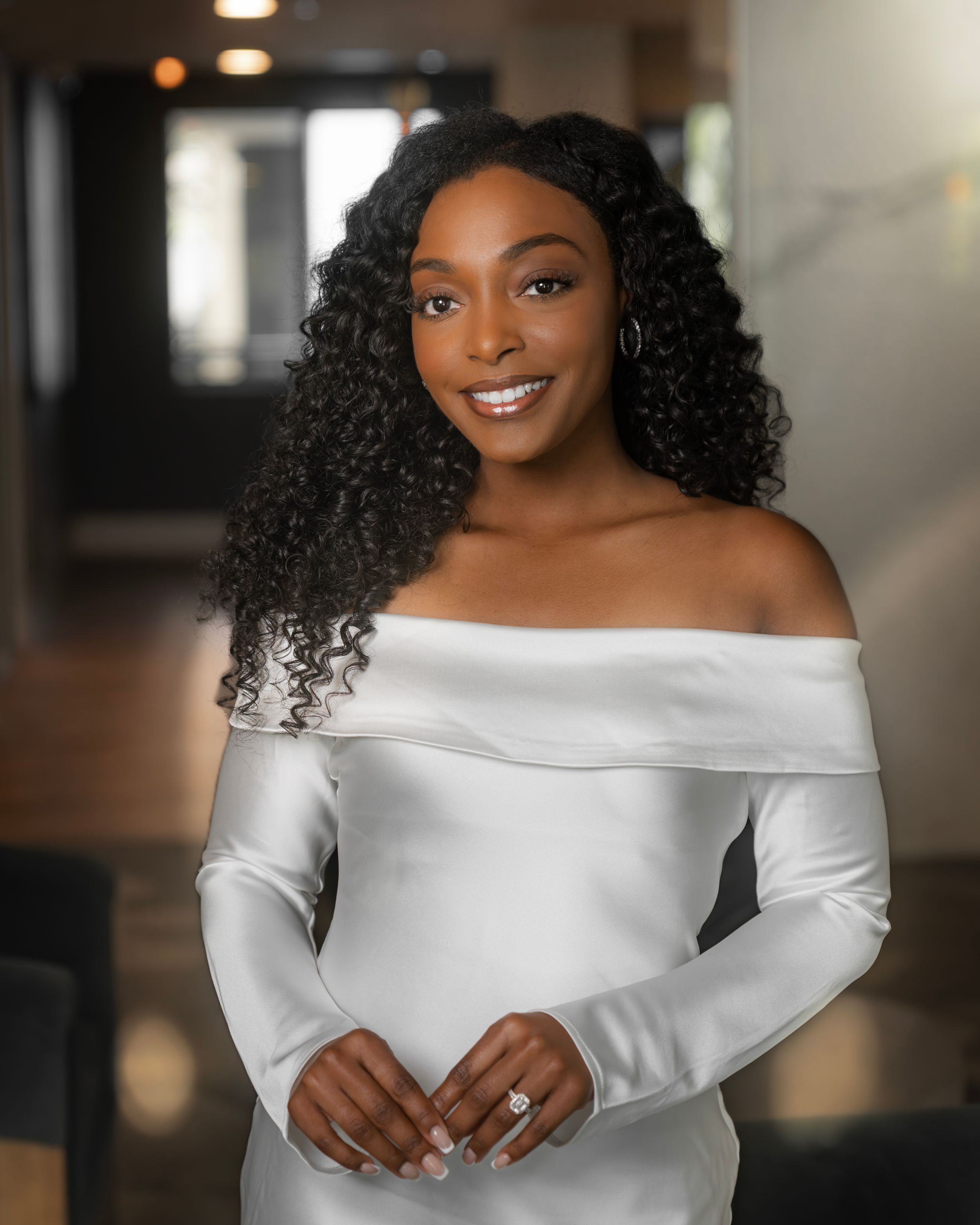 A smiling woman in an elegant white off-shoulder satin gown with curly hair and hoop earrings poses indoors.
