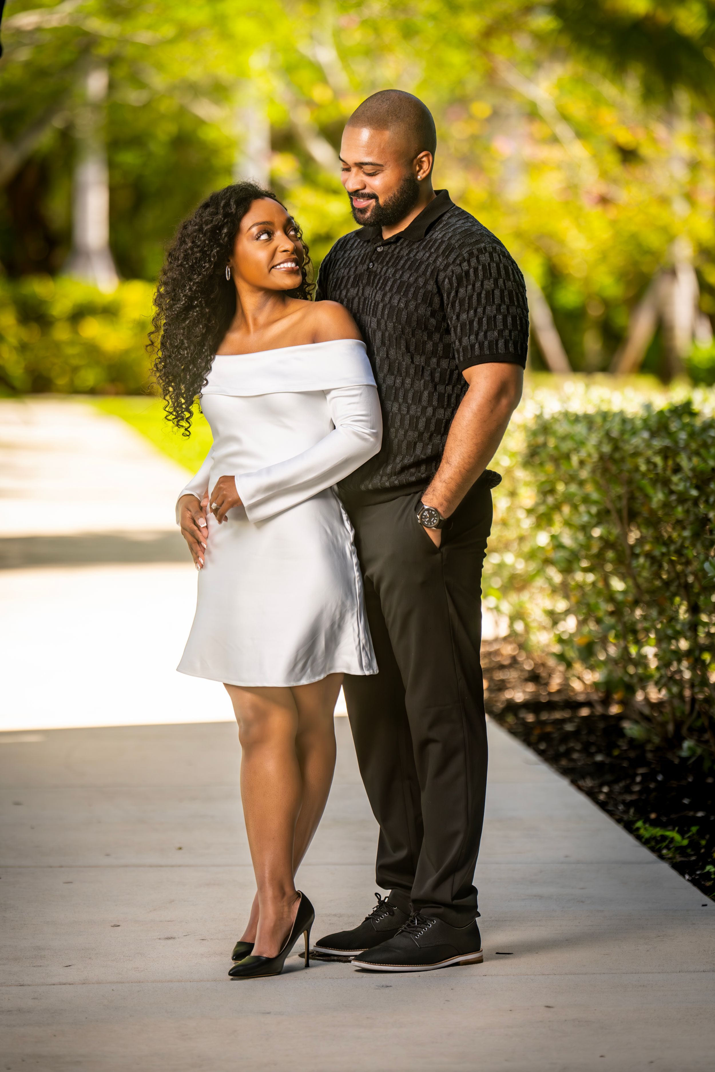 A stylish Black couple poses outdoors, woman in white off-shoulder dress, man in black shirt, surrounded by lush greenery.