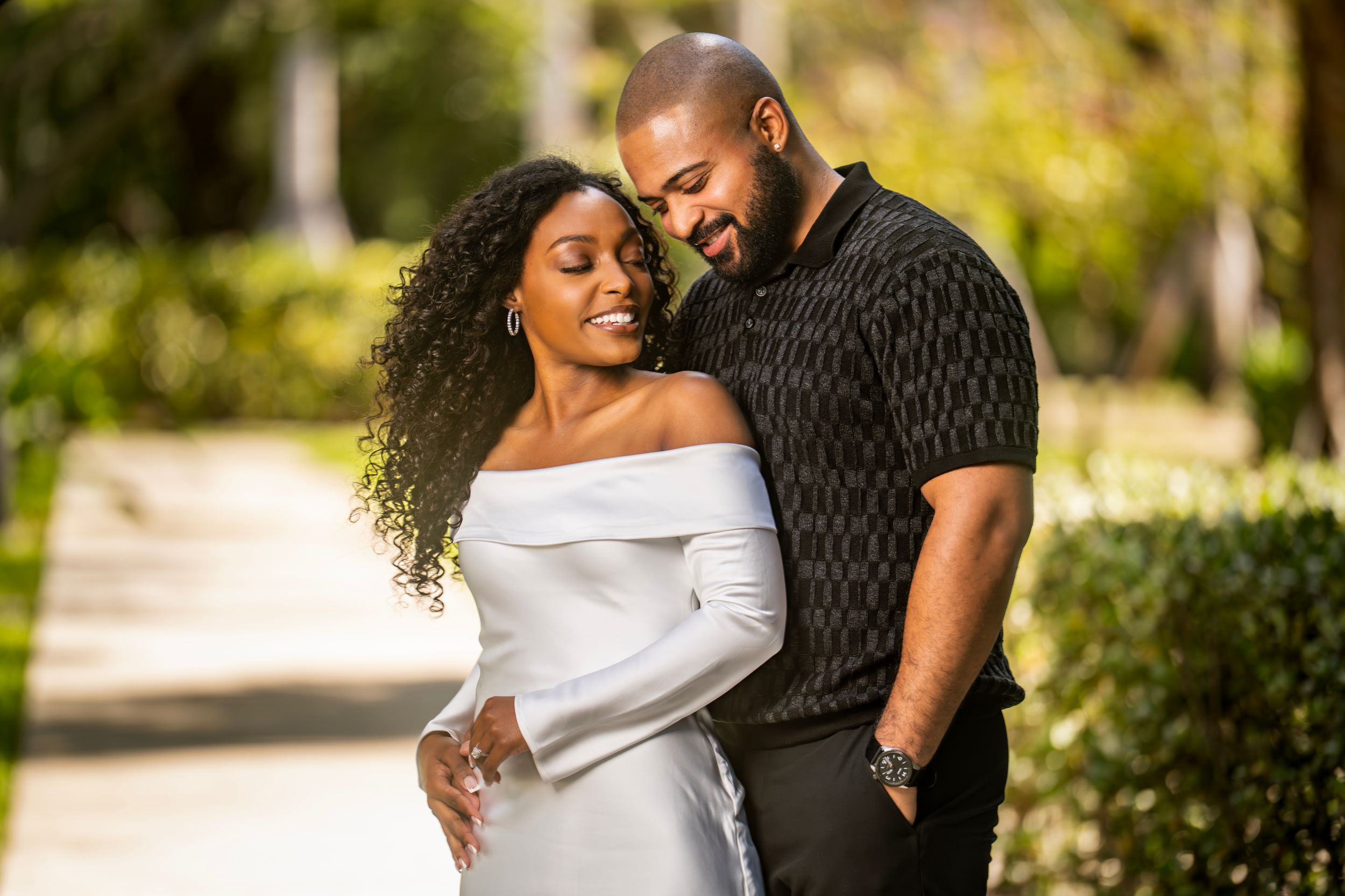 A couple embraces tenderly outdoors, woman in white off-shoulder dress, man in black shirt, surrounded by lush greenery.