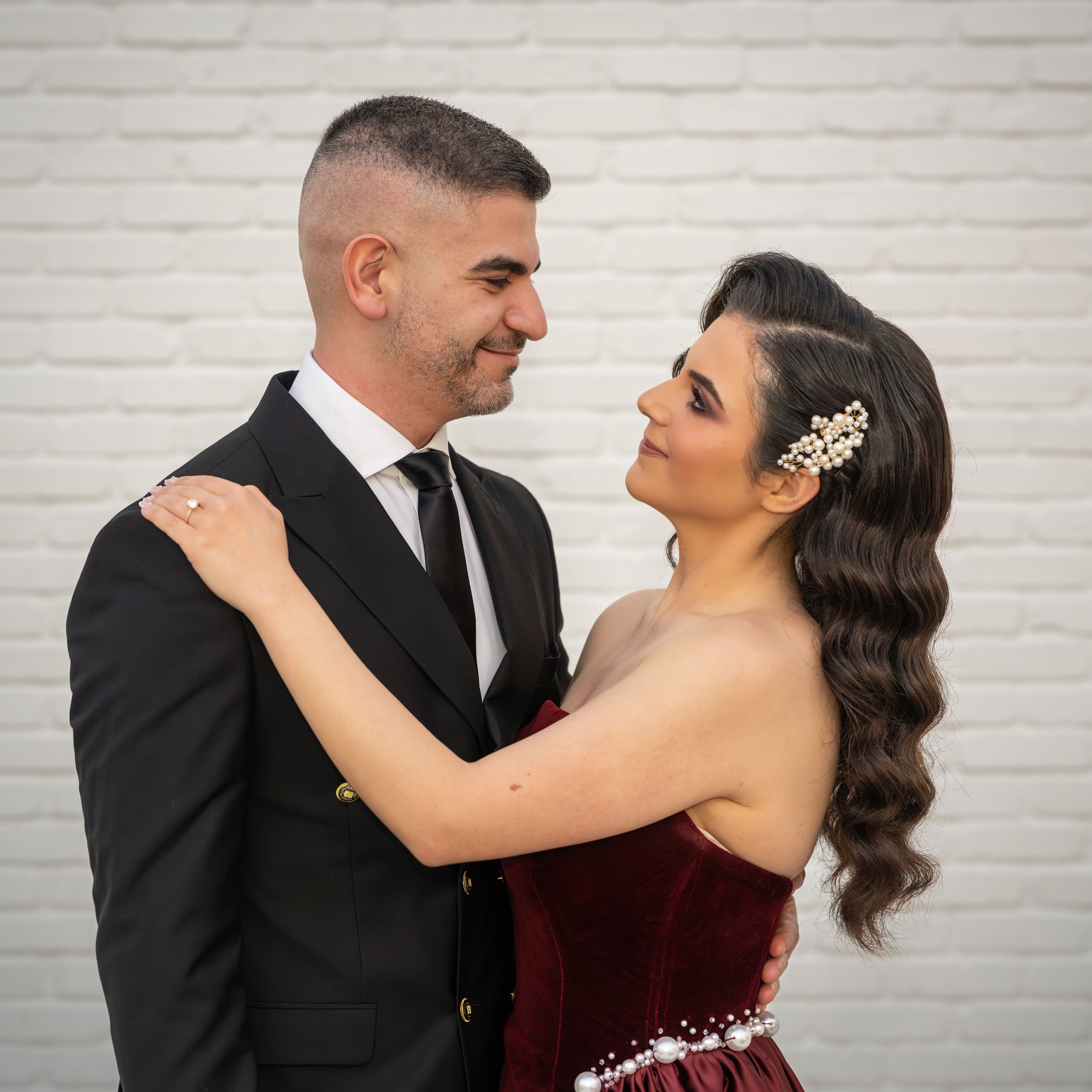 Romantic couple in formal attire share tender moments against white brick wall in wedding photo series.
