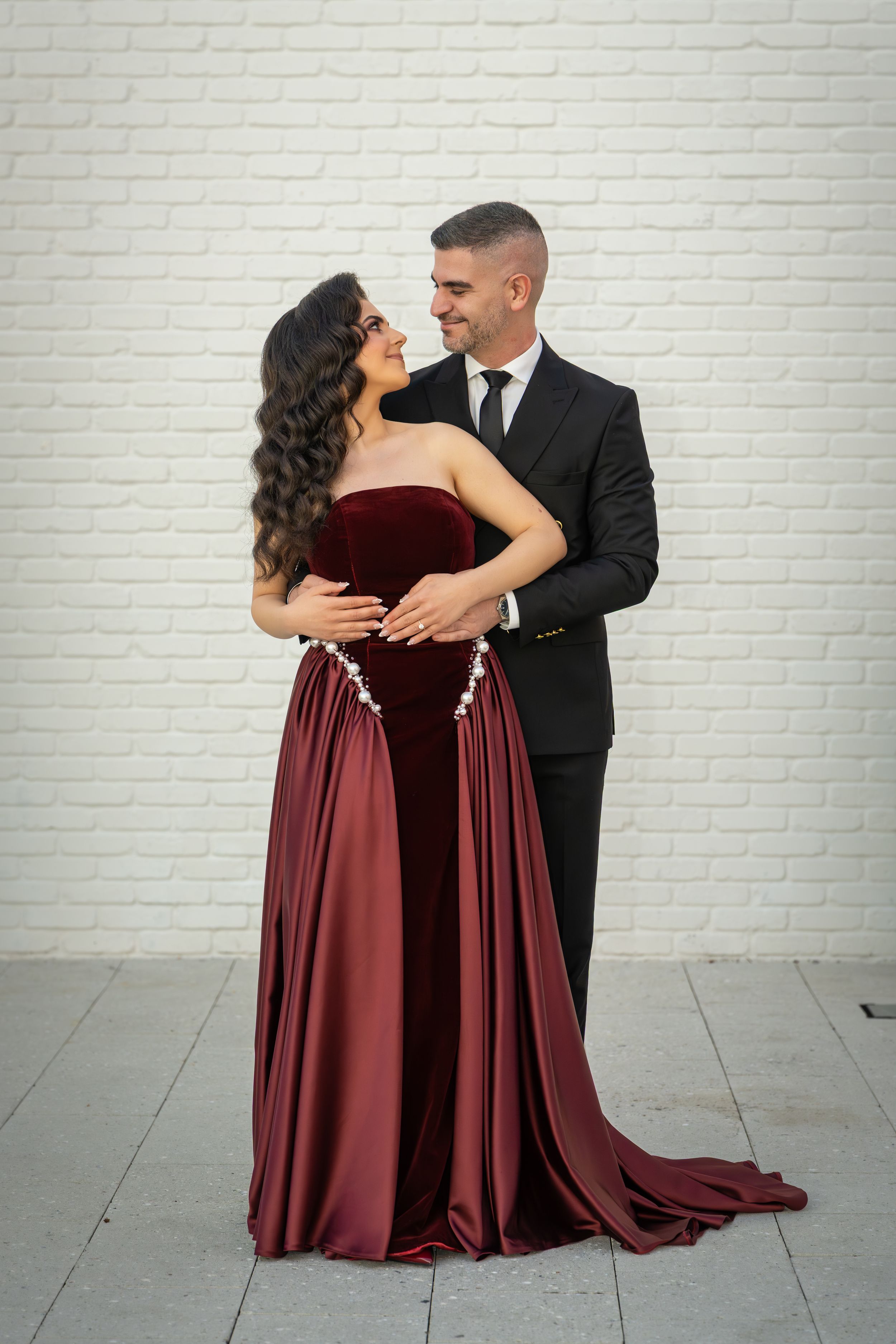 Elegant couple in formal attire dancing against a white brick wall backdrop.