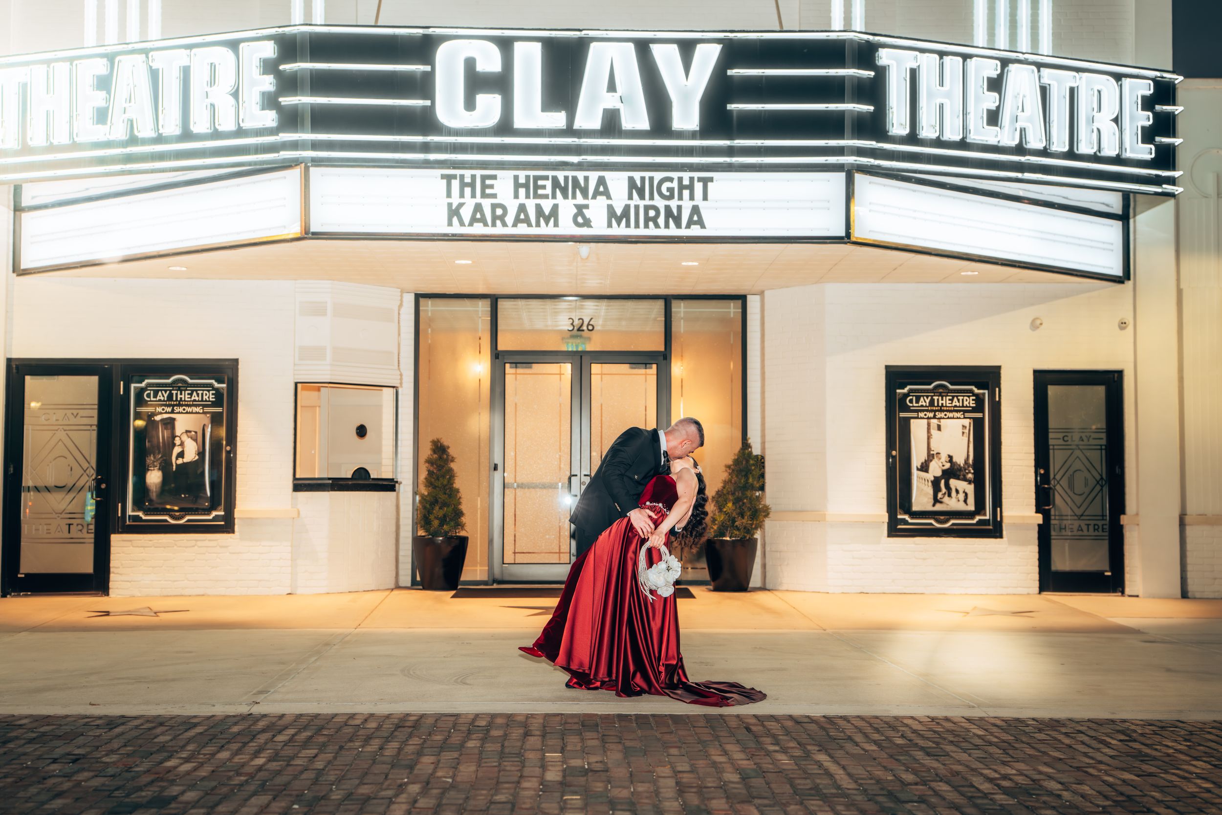 Couple embraces in front of Clay Theatre marquee displaying The Henna Night for Karam and Mirna at night.