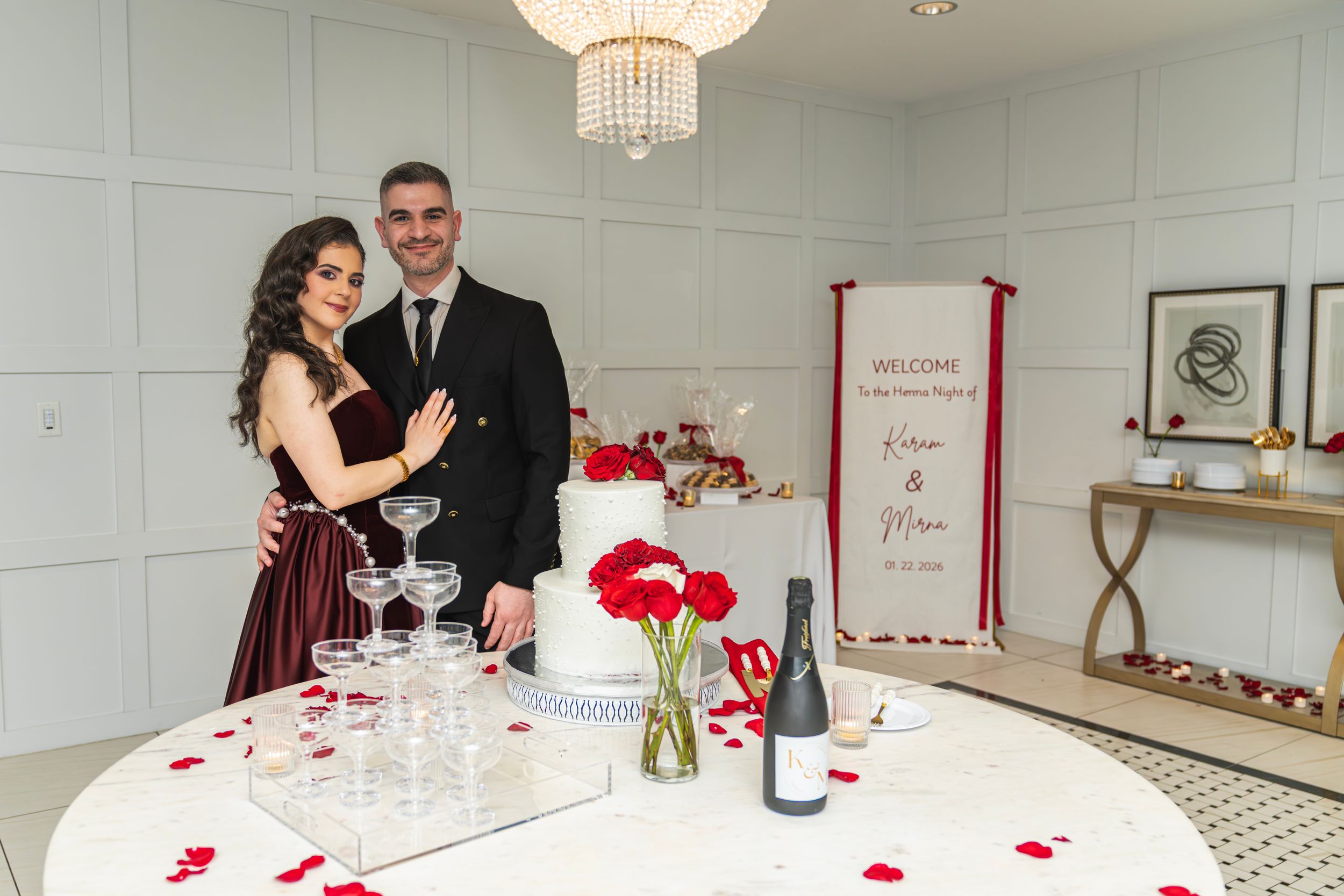Couple posing by wedding cake table with champagne tower, red roses, and welcome sign in elegant white venue.