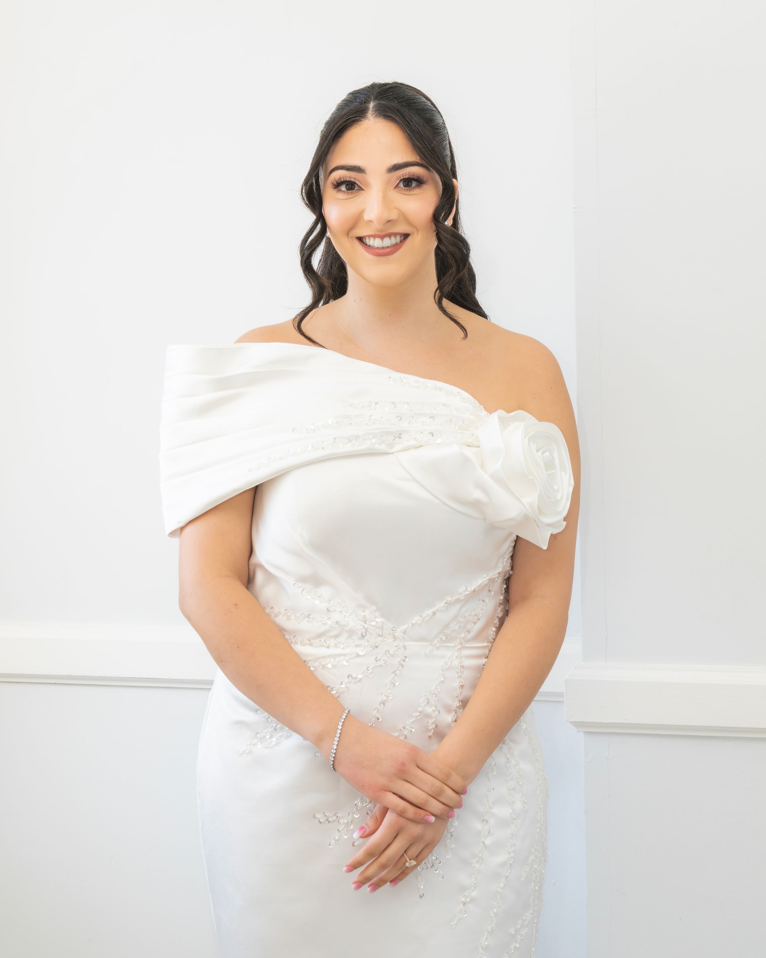 A bride poses in an elegant off-shoulder white wedding gown with delicate lace details against a light background.
