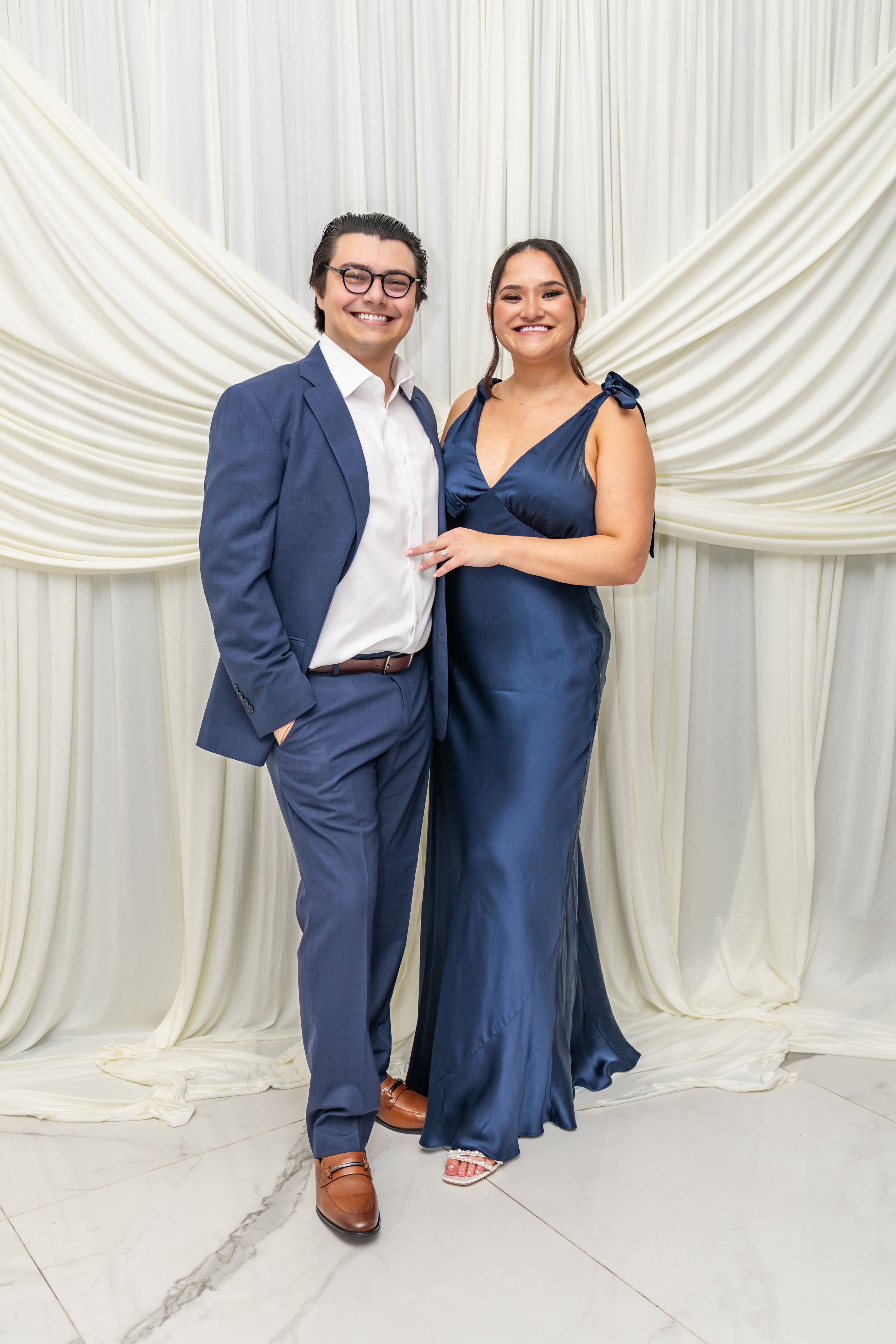 Couple in matching navy blue formal attire posing together in front of elegant white draped backdrop.