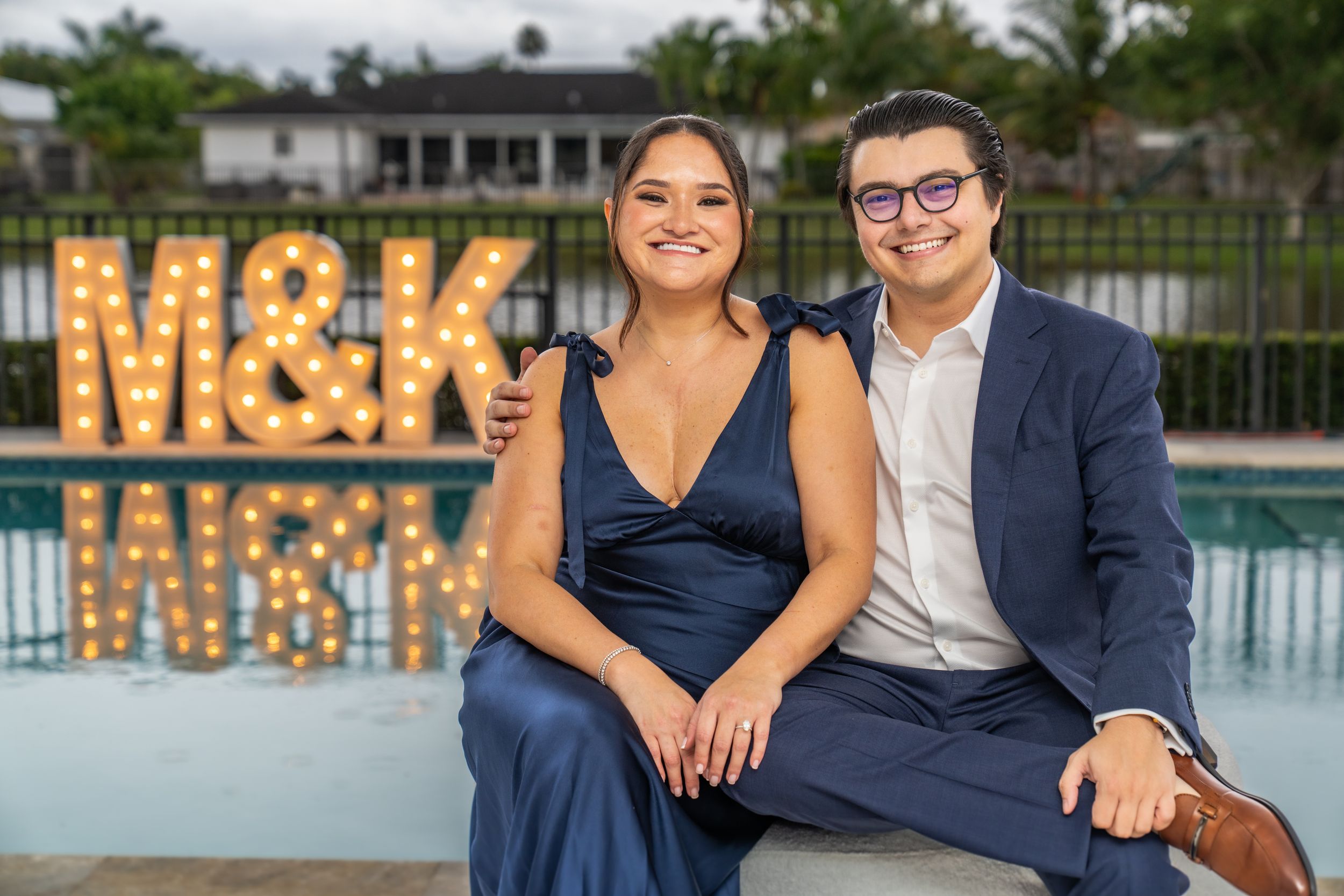 A couple in formal navy attire poses together at an outdoor event with illuminated marquee letters in the background.