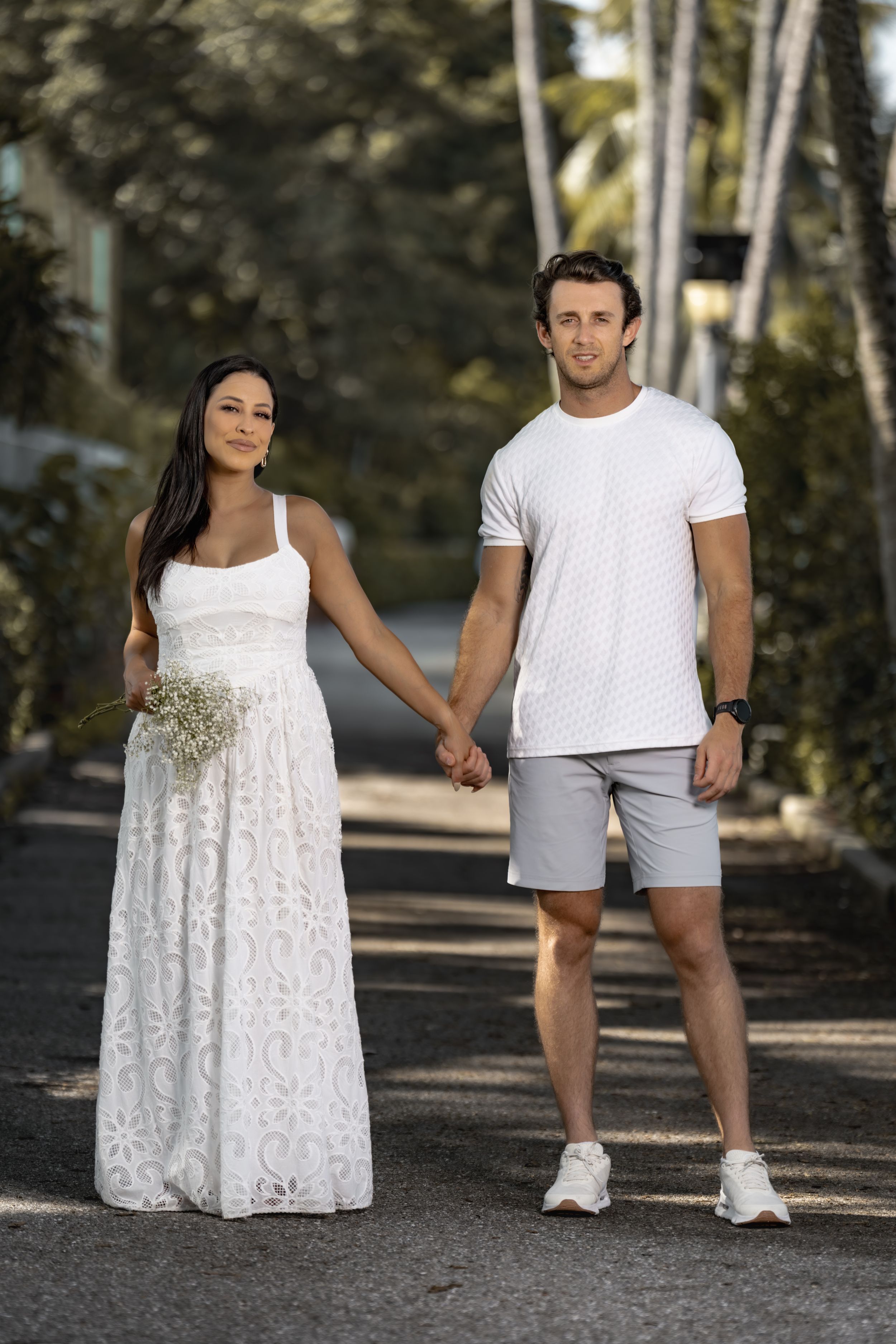 A couple walking on a path wearing white outfits, with trees in the background creating a romantic outdoor setting.