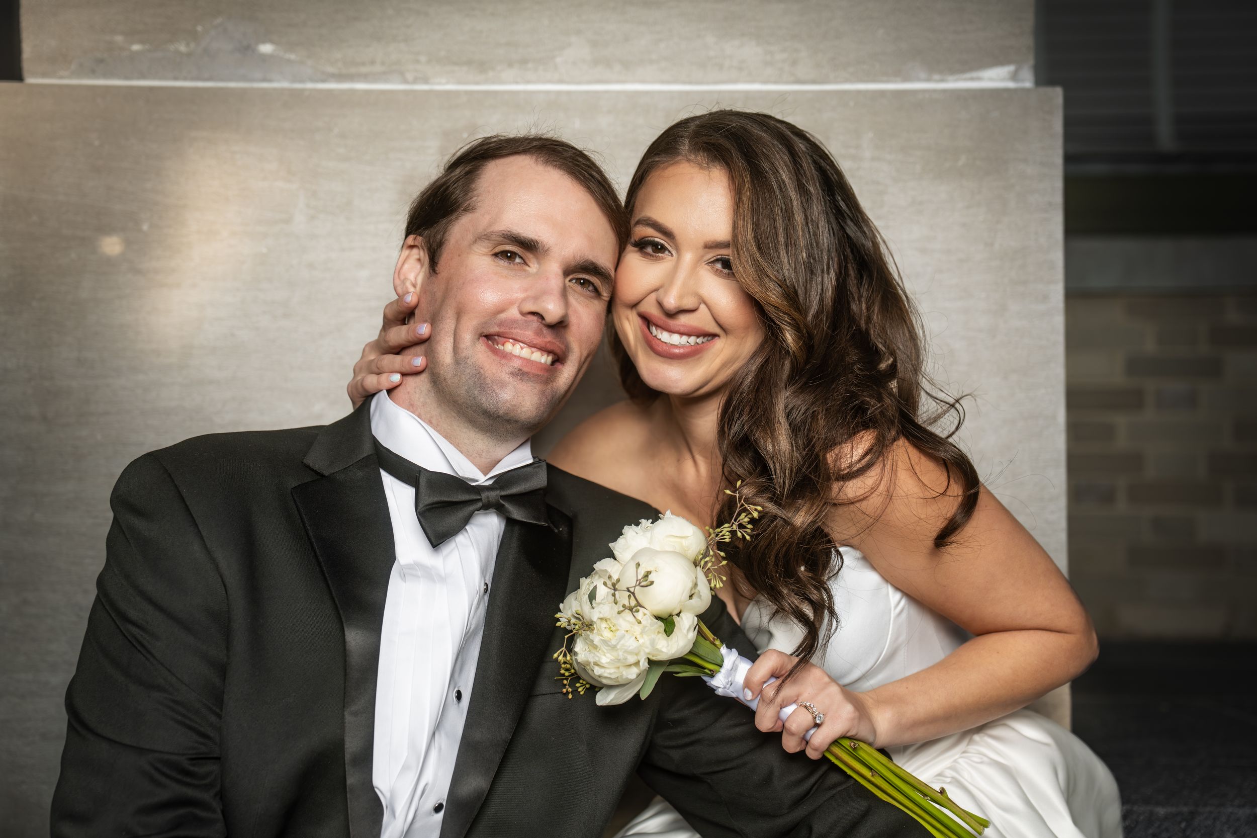 A smiling bride and groom pose together on their wedding day, she holds a white bouquet while wearing a strapless gown.