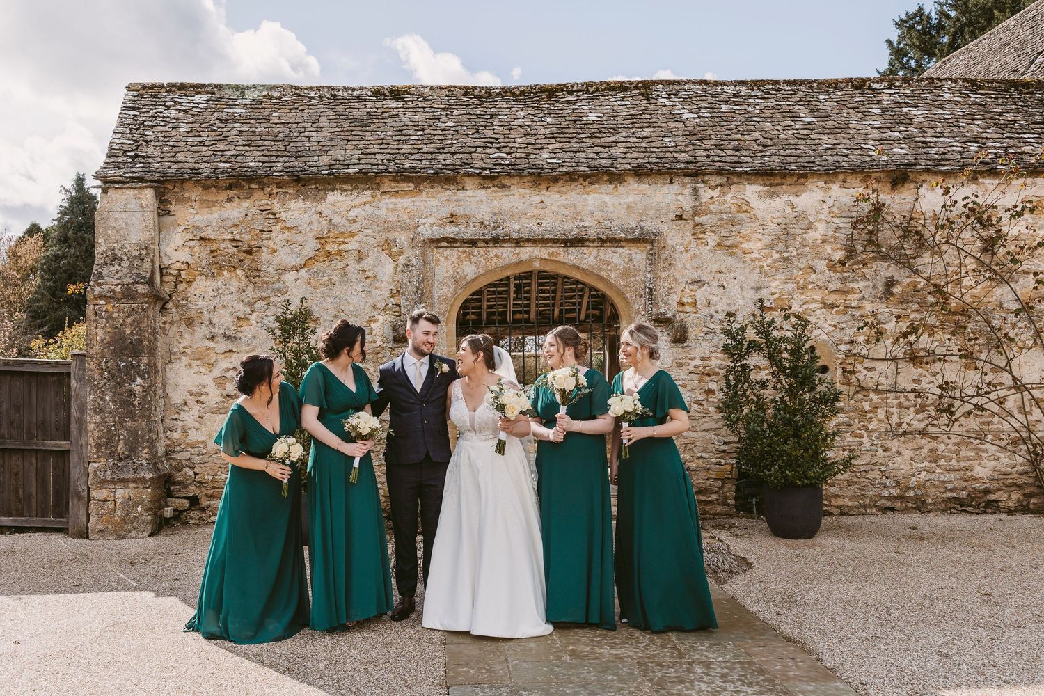 Wedding party wearing emerald green dresses poses together at a rustic stone venue for outdoor photos at Caswell House
