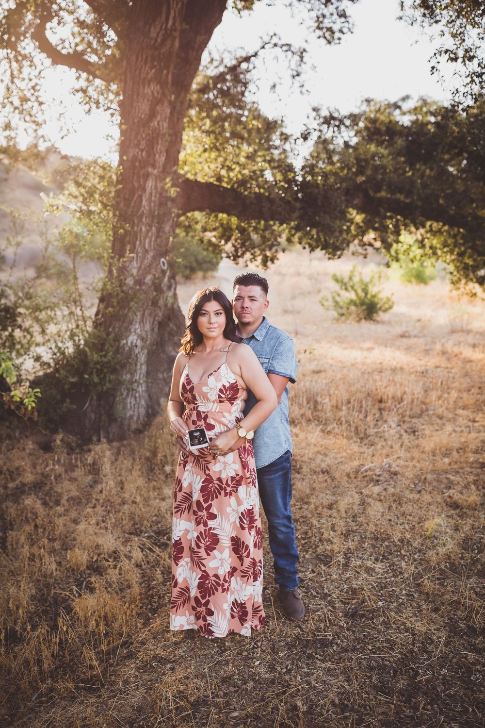 A couple shares a romantic moment in a sunlit field with golden grass and trees in the background.