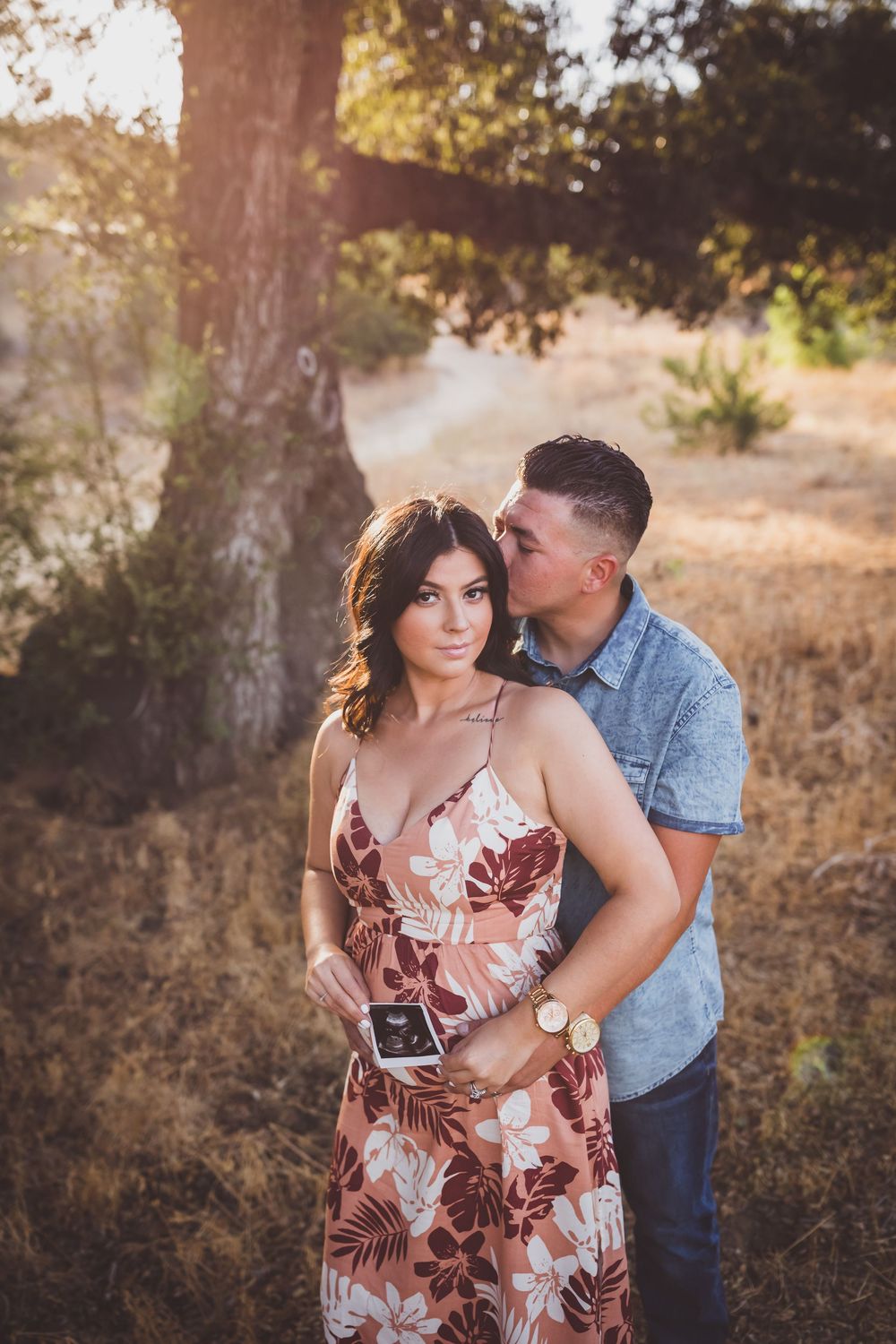 A couple embraces while holding an ultrasound photo in a scenic outdoor setting during golden hour.