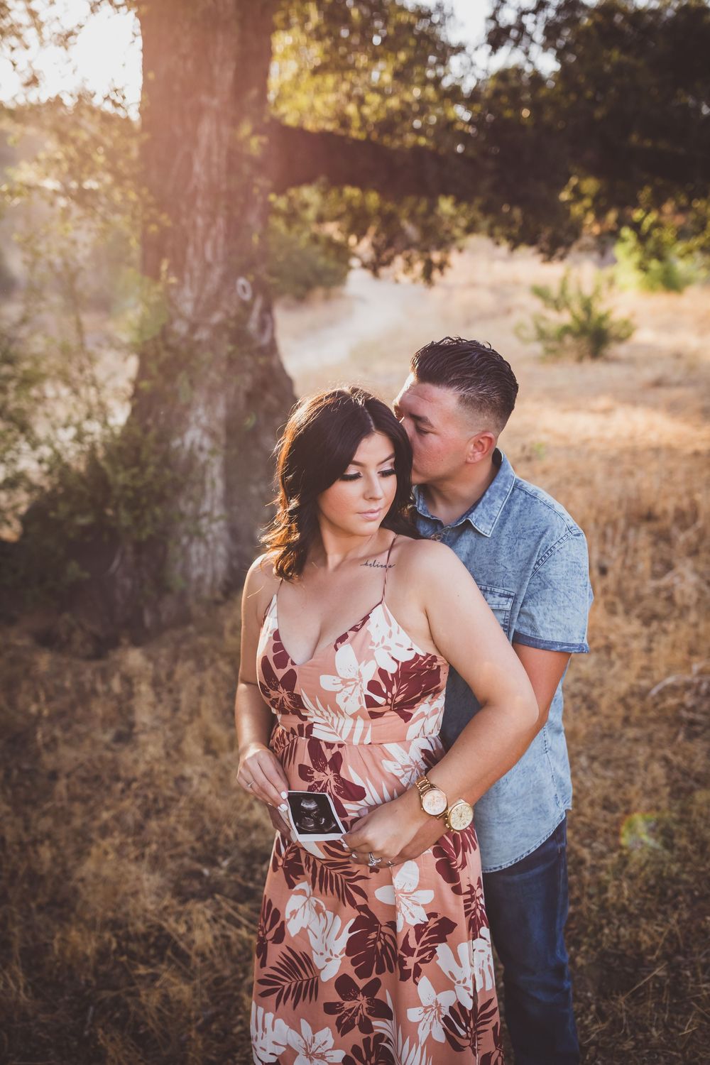 A couple embraces in a romantic outdoor photoshoot during golden hour among trees and tall grass.