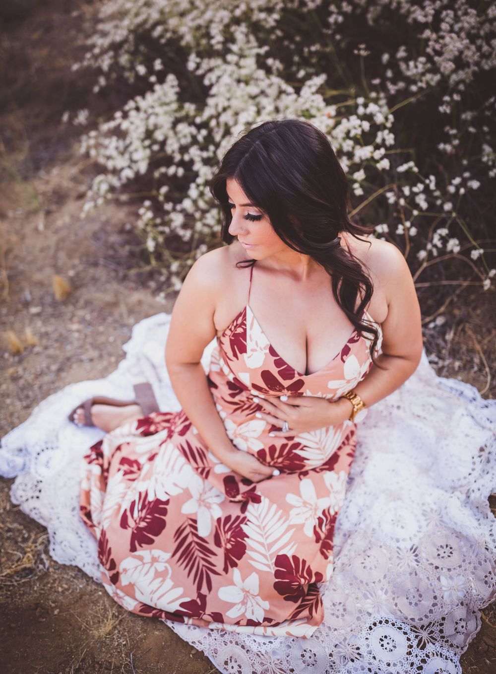 A woman in a floral pink and burgundy dress poses gracefully on white lace fabric in an outdoor setting.