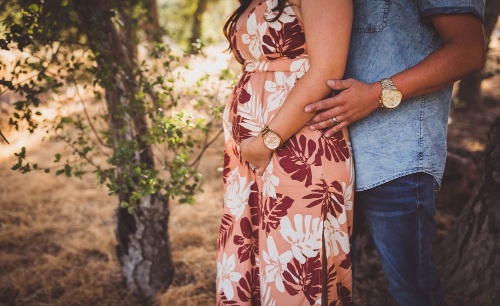 A couple in casual attire embraces tenderly during an outdoor photoshoot in a sun-dappled forest setting.