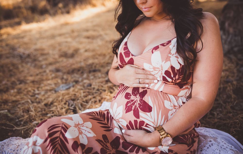 A woman in a floral dress sits on the ground during a sunset photoshoot with soft natural lighting.