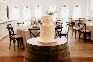 White  wedding cake displayed on rustic wooden barrel in elegant reception venue with arched windows by Wedding photographer Serene Coleman