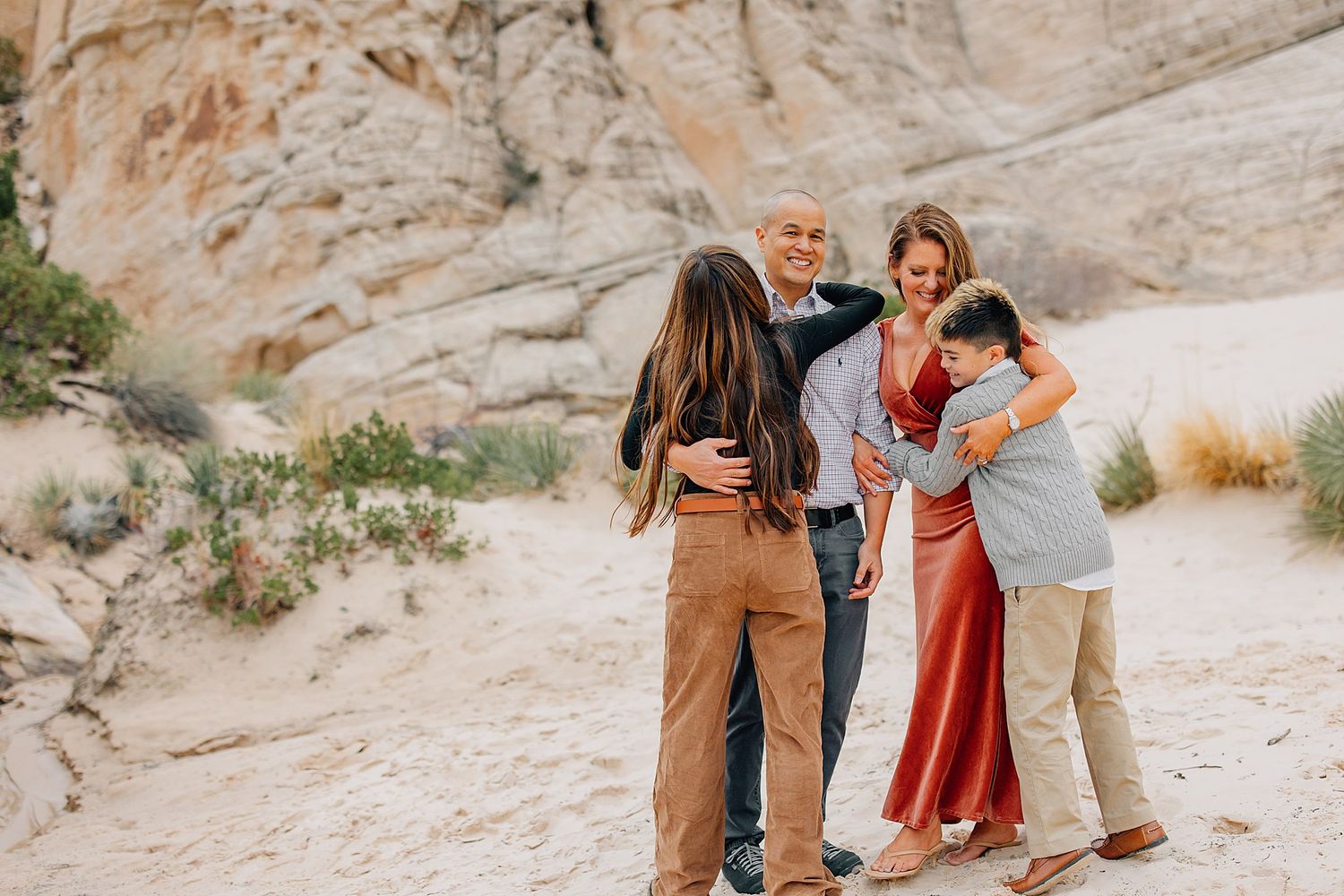 Family embracing and walking together on rocky desert terrain wearing coordinated rust and neutral colored outfits.