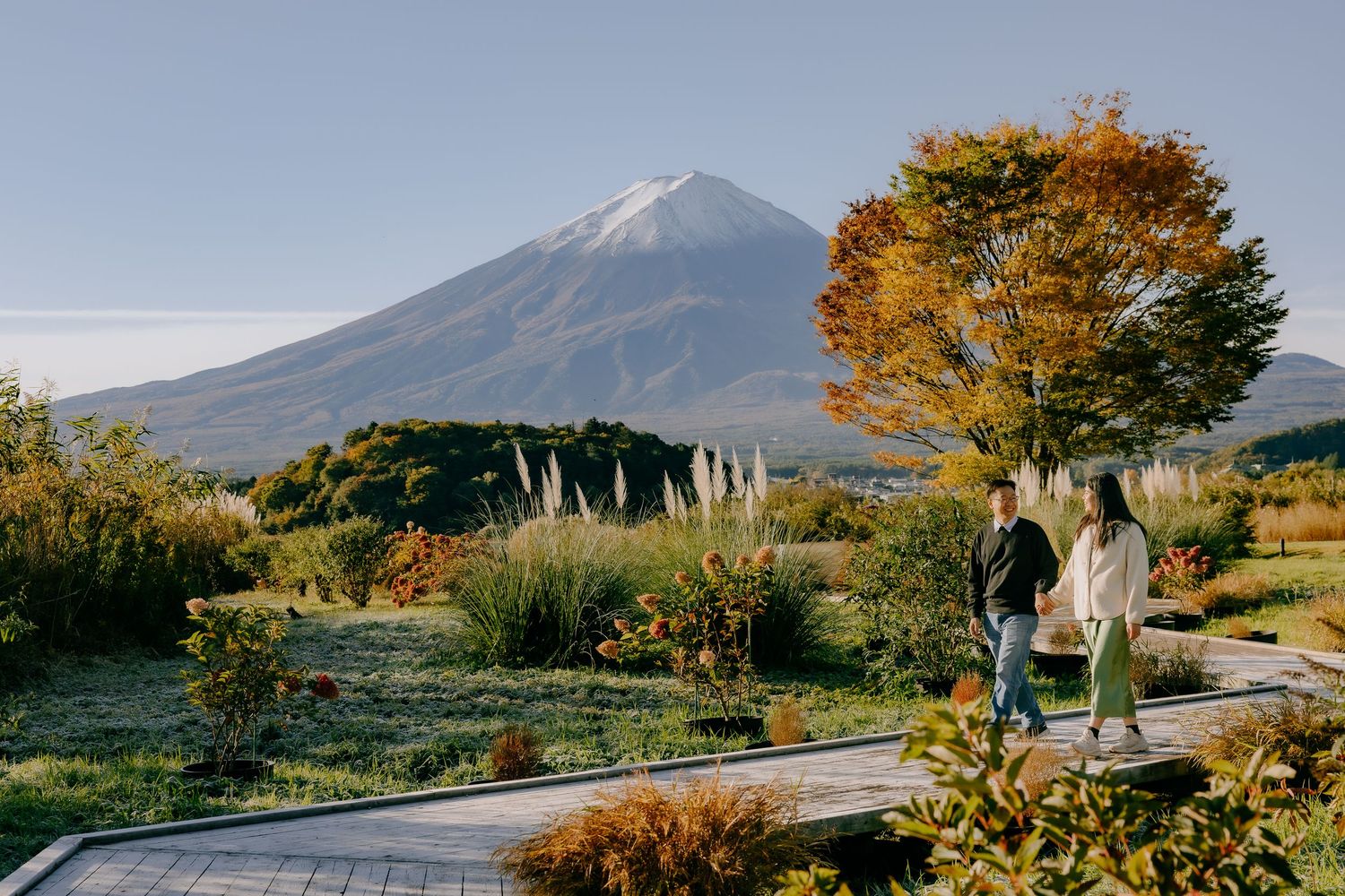 Two people walking on a pathway surrounded by autumn foliage with Mount Fuji rising majestically in the background.