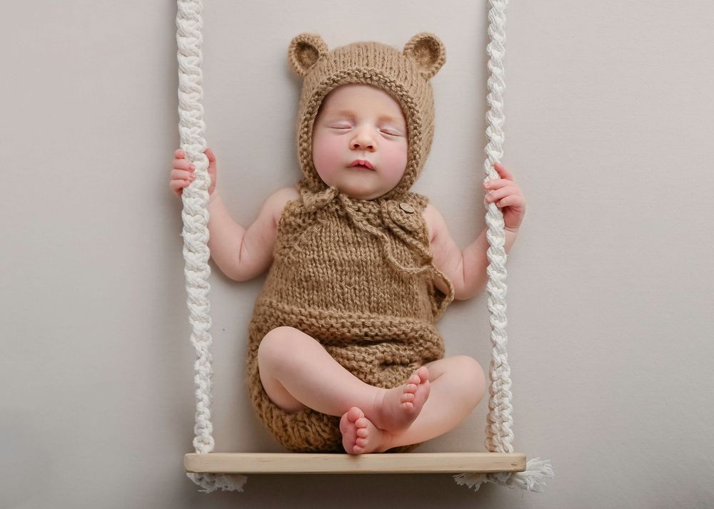 Baby wearing knitted brown bear outfit sits contentedly on a white rope swing against gray background.