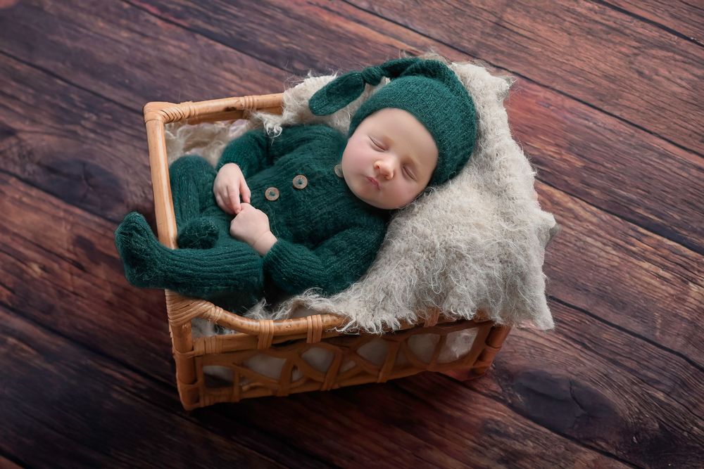 Baby in teal sweater rests peacefully on fluffy white blanket in wooden basket on dark wood floor.