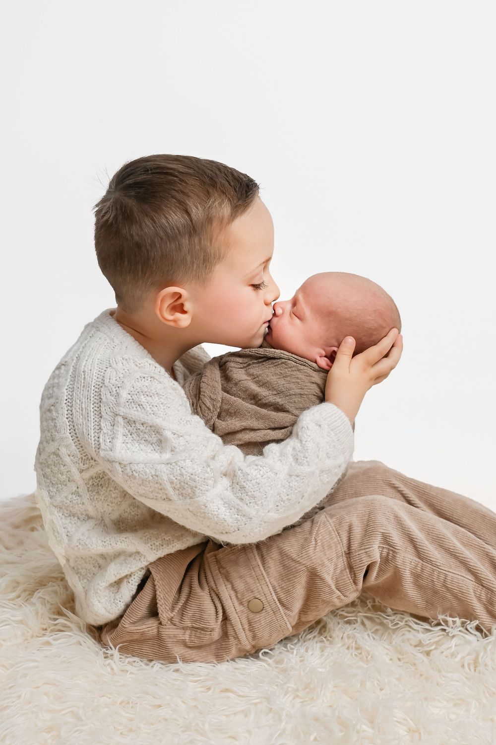 Two siblings in white and beige clothing share a tender moment lying on a fluffy white rug.