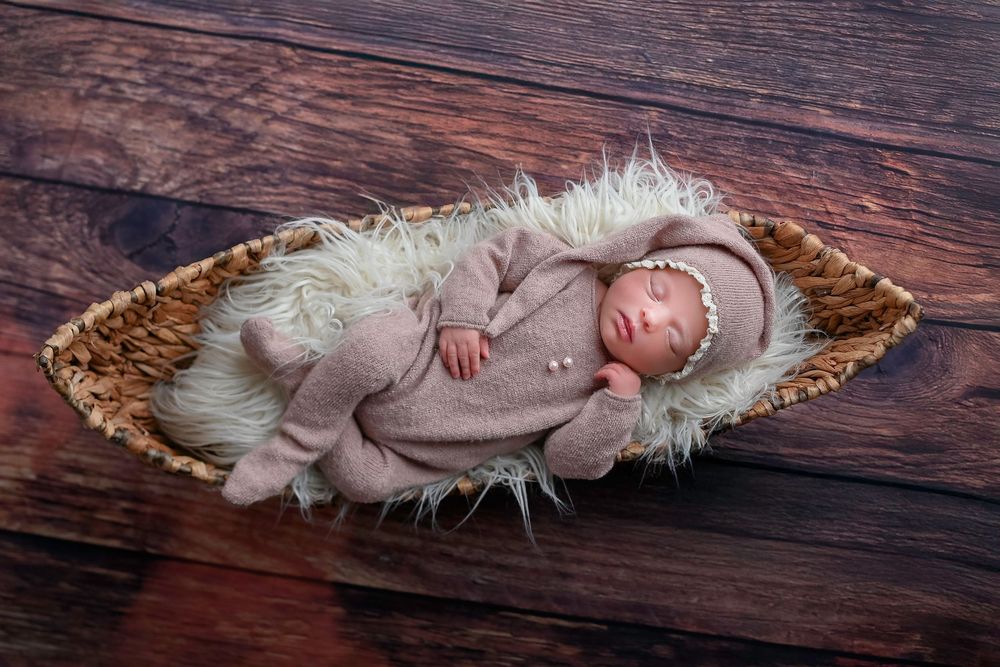 A sleeping newborn wrapped in a soft gray swaddle rests peacefully in a rustic wooden basket with white fur padding.