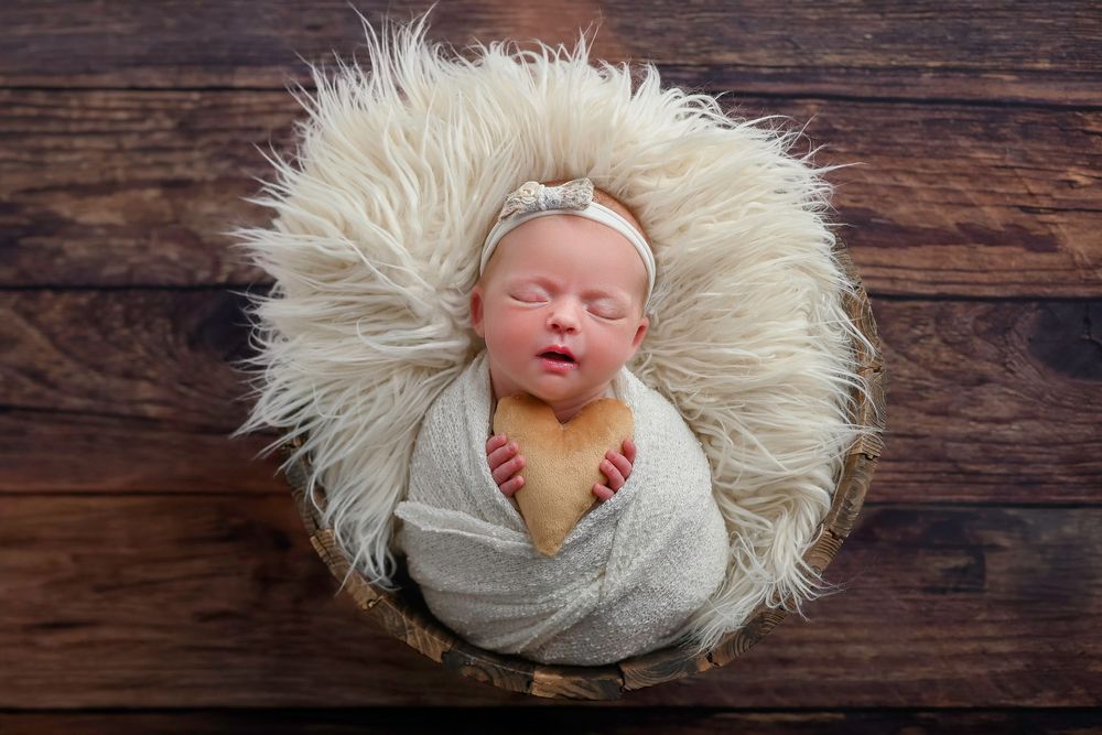 A sleeping newborn wrapped in a gray swaddle blanket lies on a white furry rug against a rustic wooden backdrop.