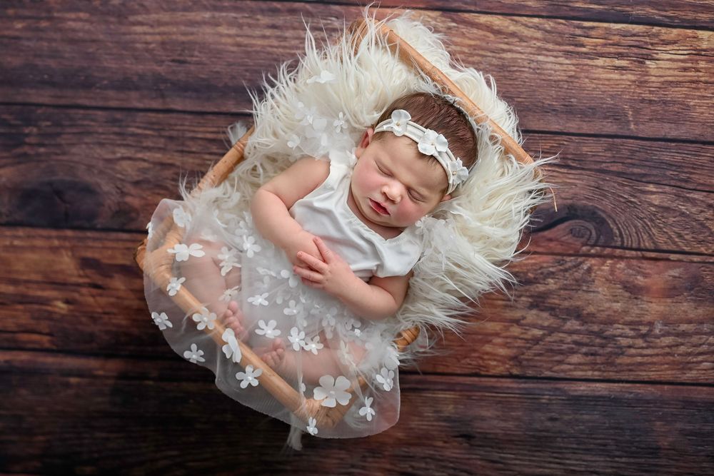 Sleeping newborn baby wearing a white lace dress lies on soft fur blanket against rustic wooden background.