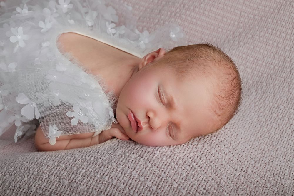 A sleeping newborn baby in a white lace wrap lies on a soft textured gray backdrop.