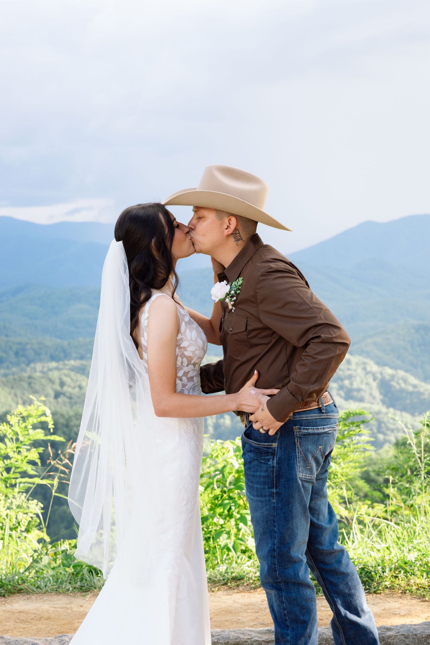 A couple in western attire shares a romantic kiss against a scenic mountain backdrop with a bride in white dress and veil.
