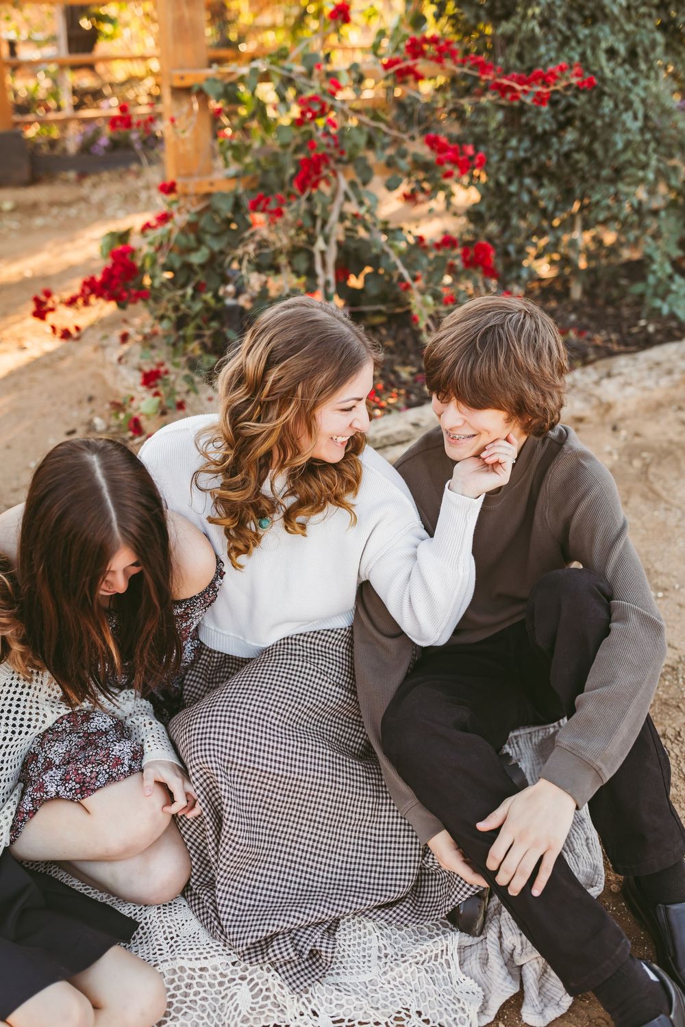 Three friends share a candid moment of laughter while sitting together outdoors with red flowers in the background.