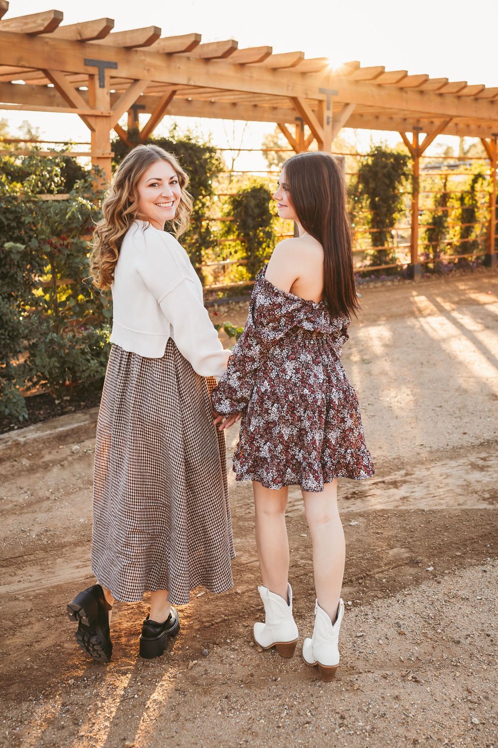 Two people in fall outfits pose playfully under a garden pergola with warm sunlight streaming through.