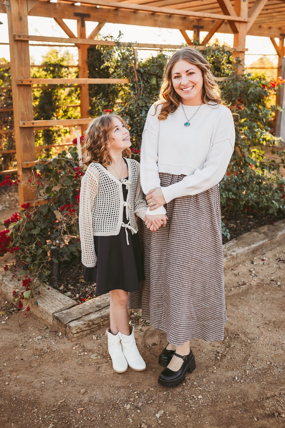Two people in coordinating black and white outfits stand together under a wooden pergola with blooming roses.