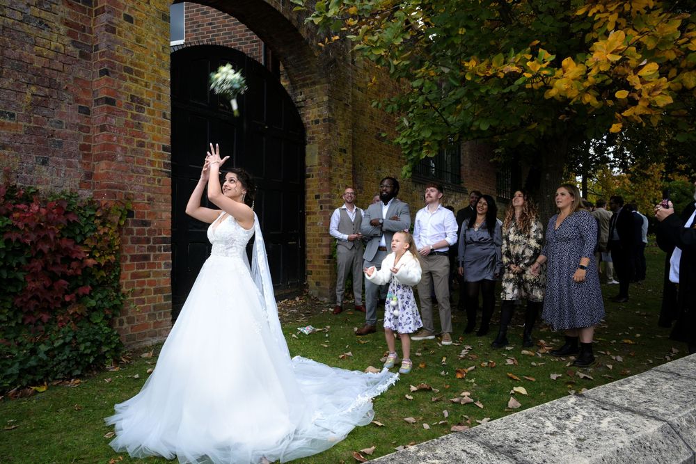 A bride in a flowing white gown tosses her bouquet at an outdoor wedding ceremony while guests watch from nearby.