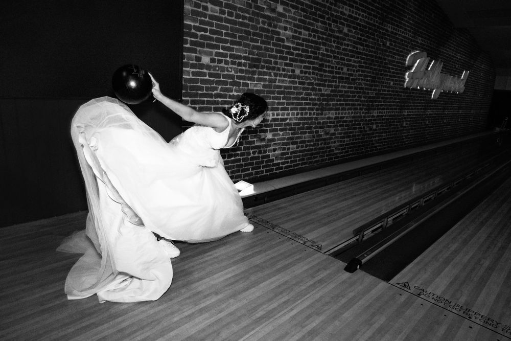 A person in a white protective suit crouches on a bowling lane at night, taking a low-angle photograph.