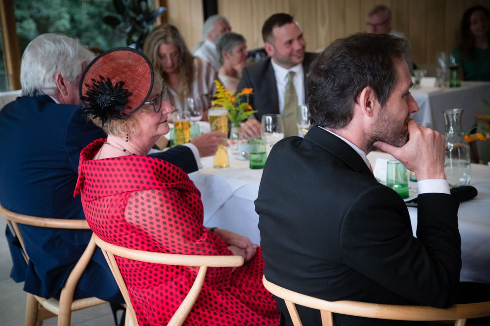 Wedding guests in formal attire sit at a reception table with someone wearing a bright red dress and fascinator hat. WALES