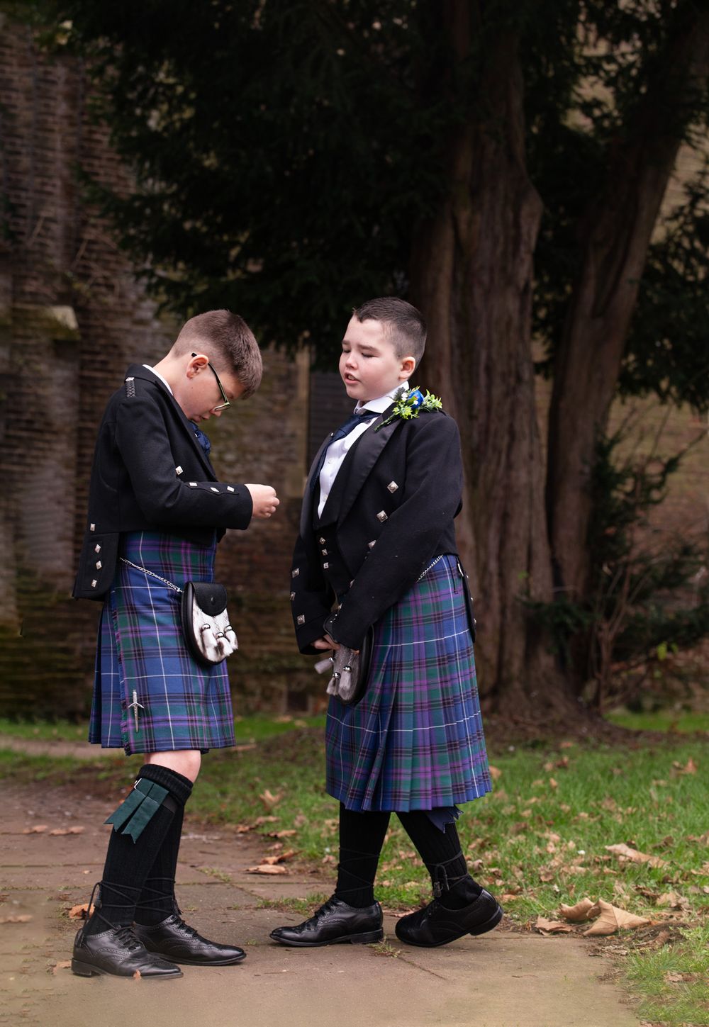 Two people in traditional Scottish kilts and formal attire stand together on a path near trees and a brick wall.