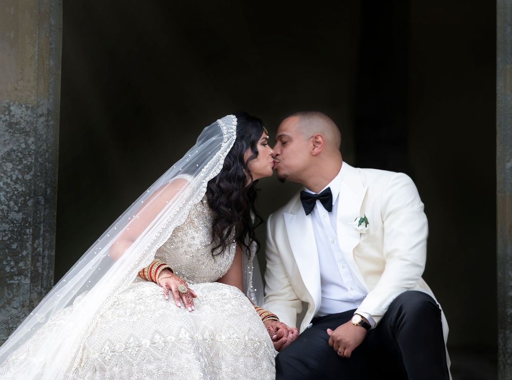 A bride in a white lace gown and groom in a white tuxedo share a romantic kiss while seated.