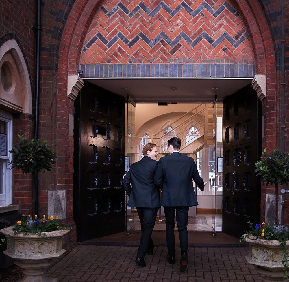 Two people in formal attire walk together through an elegant brick archway decorated with potted plants.
