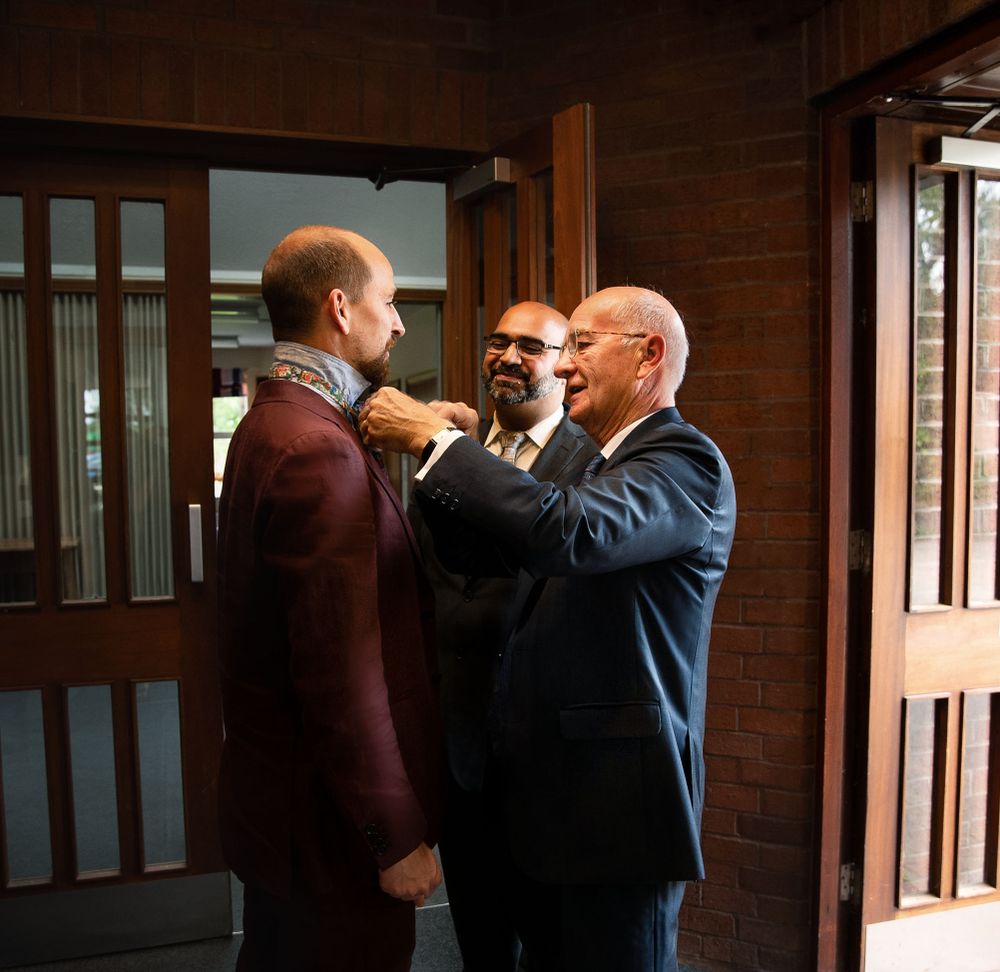 Three people stand in conversation at a doorway, wearing formal attire and engaged in a cordial interaction.