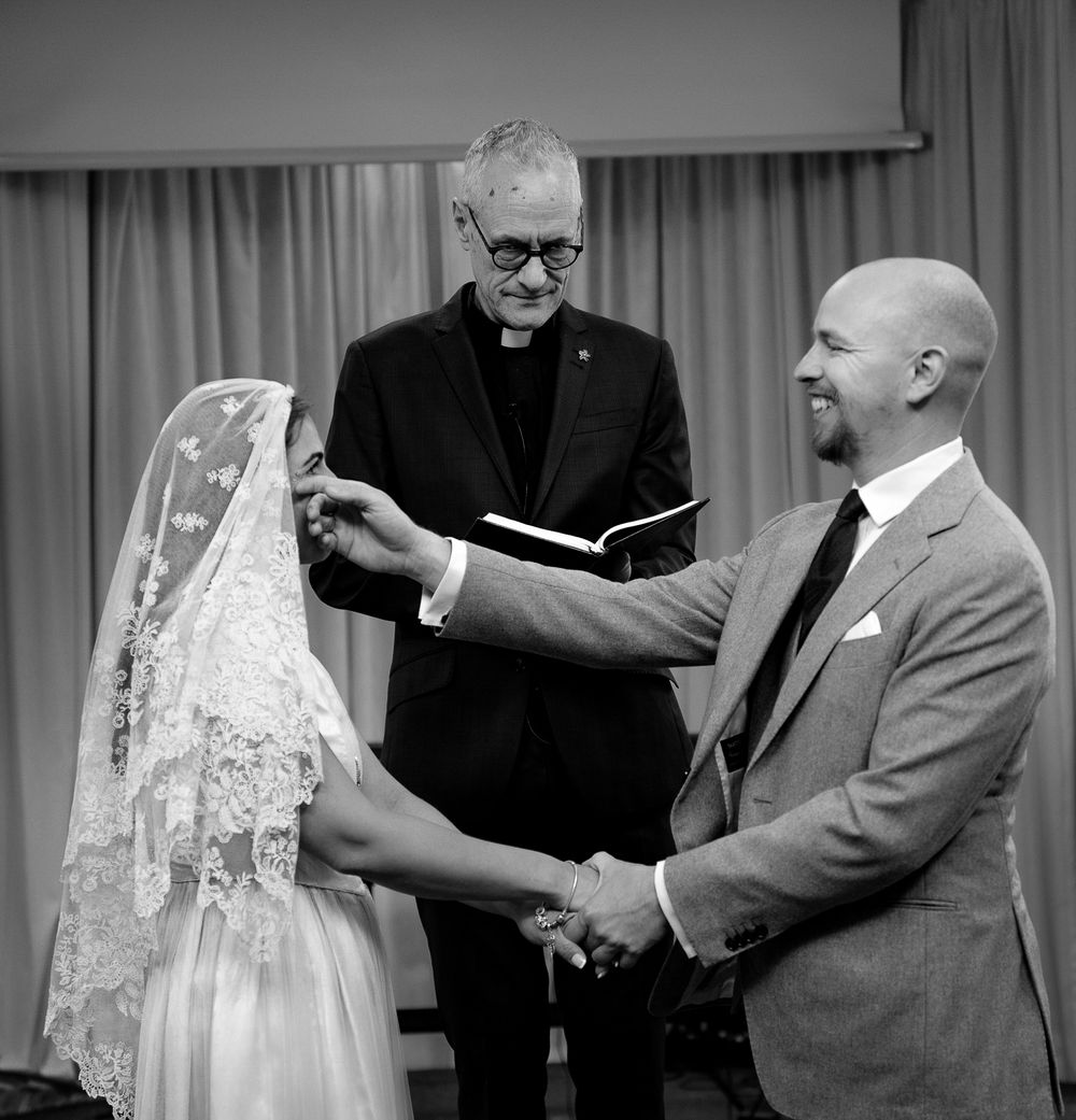 Black and white photo of a wedding ceremony with a couple holding hands as an officiant reads from a book during the service.