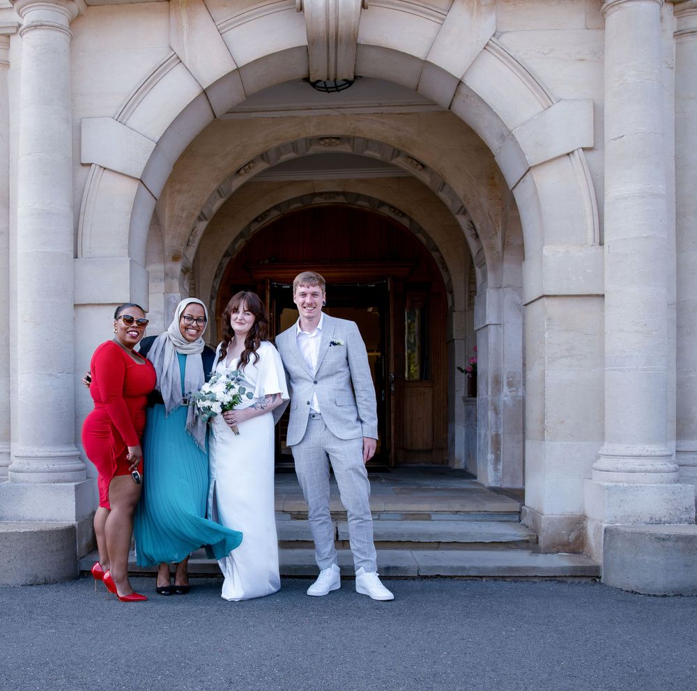 A wedding party poses beneath a grand stone archway entrance of a historic building.