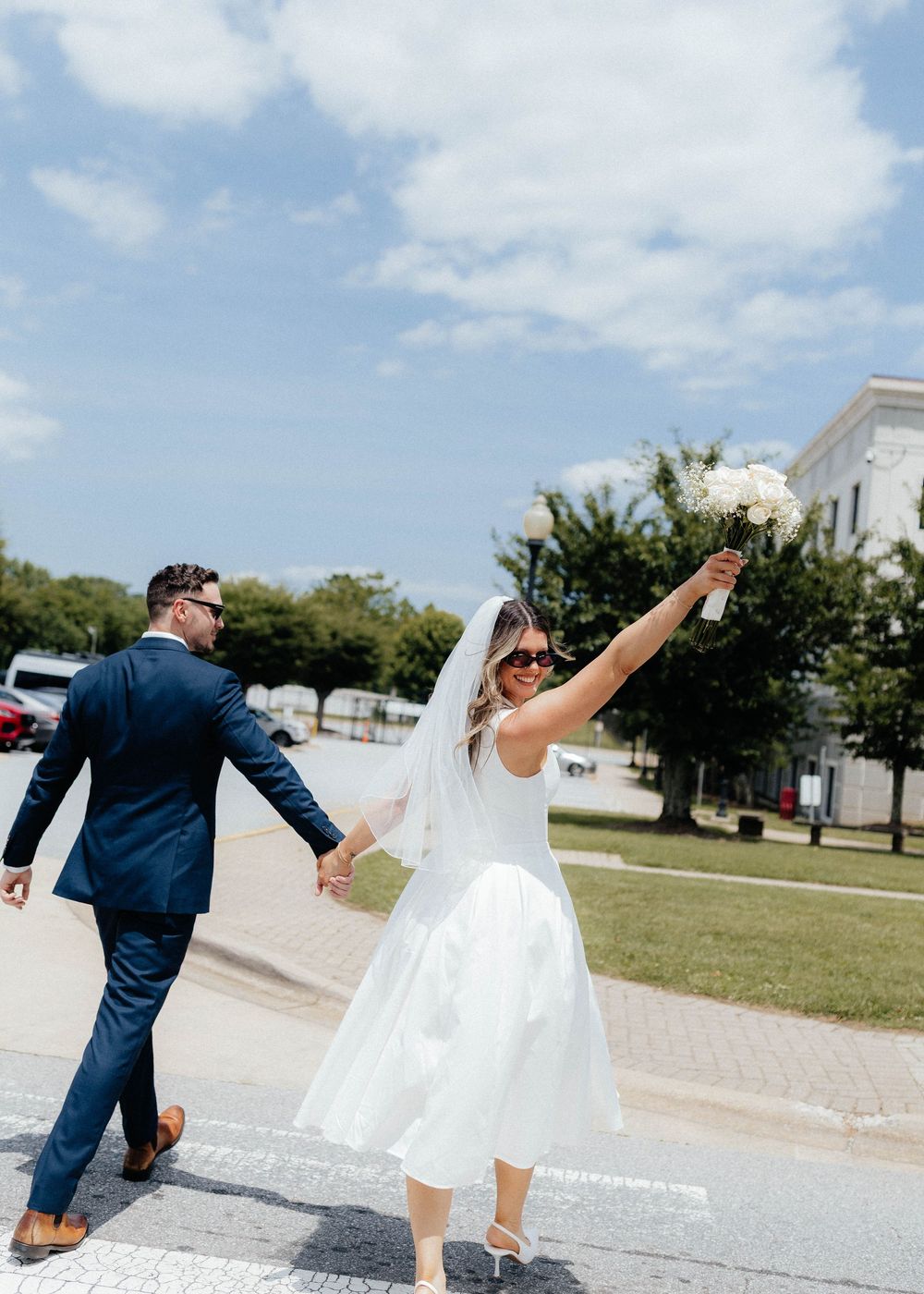 A couple in wedding attire dances playfully across a sunny outdoor walkway with white buildings in the background.