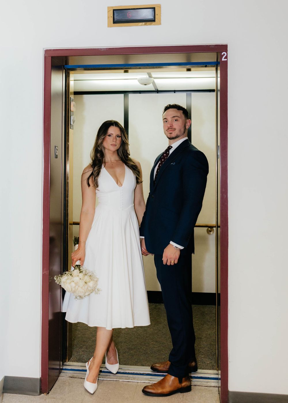 A couple in wedding attire takes a series of playful elevator photos with the bride holding a white bouquet.