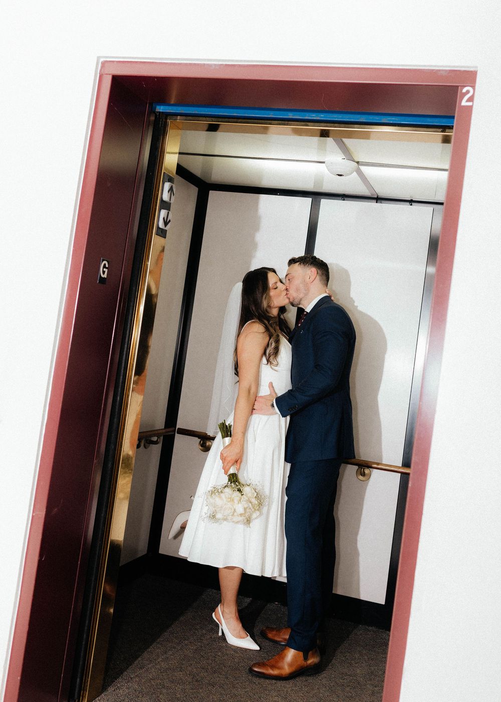 A couple shares a romantic moment in an elevator, with the bride in a white tea-length dress and groom in a navy suit.